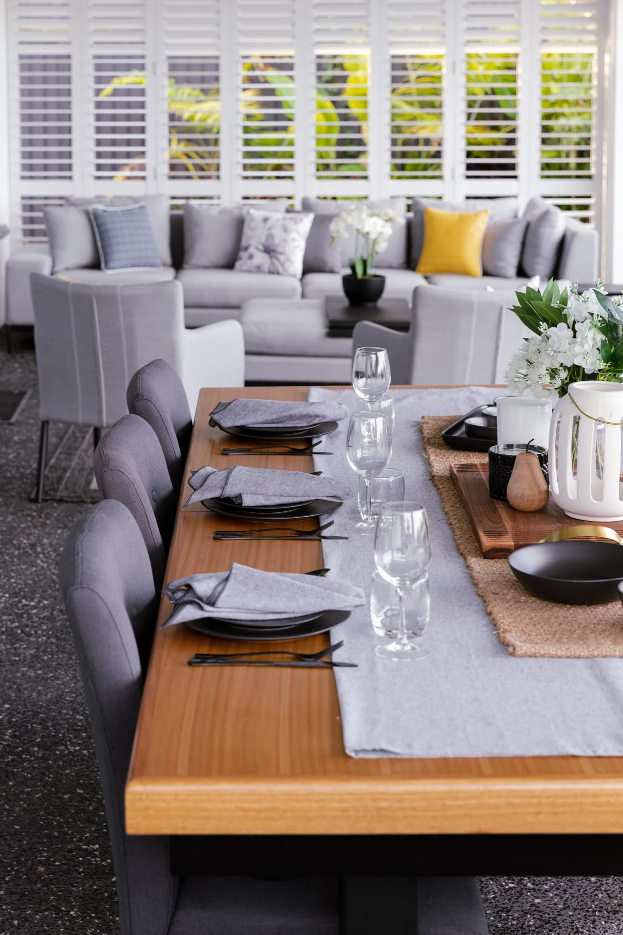 A dining table set with black plates, gray napkins, wine glasses, and silverware, with a centerpiece of white flowers and greenery in a white pitcher, on a woven placemat; in the background, a gray sofa with pillows in navy, floral, and yellow, again