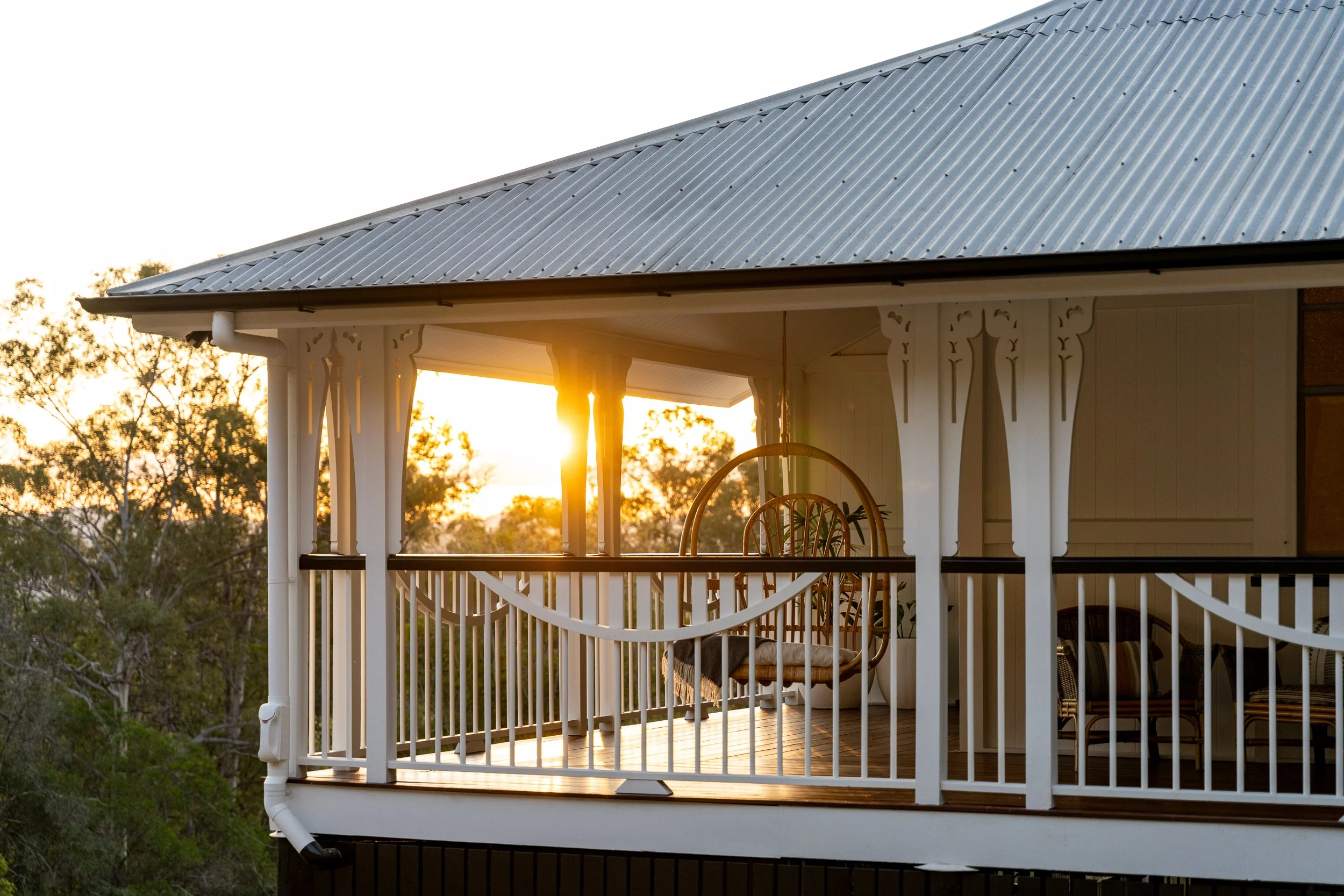 The image shows the upper part of a porch or balcony with white decorative woodwork, ledge, and railing, at sunset or sunrise, with trees in the background and the sky clear.