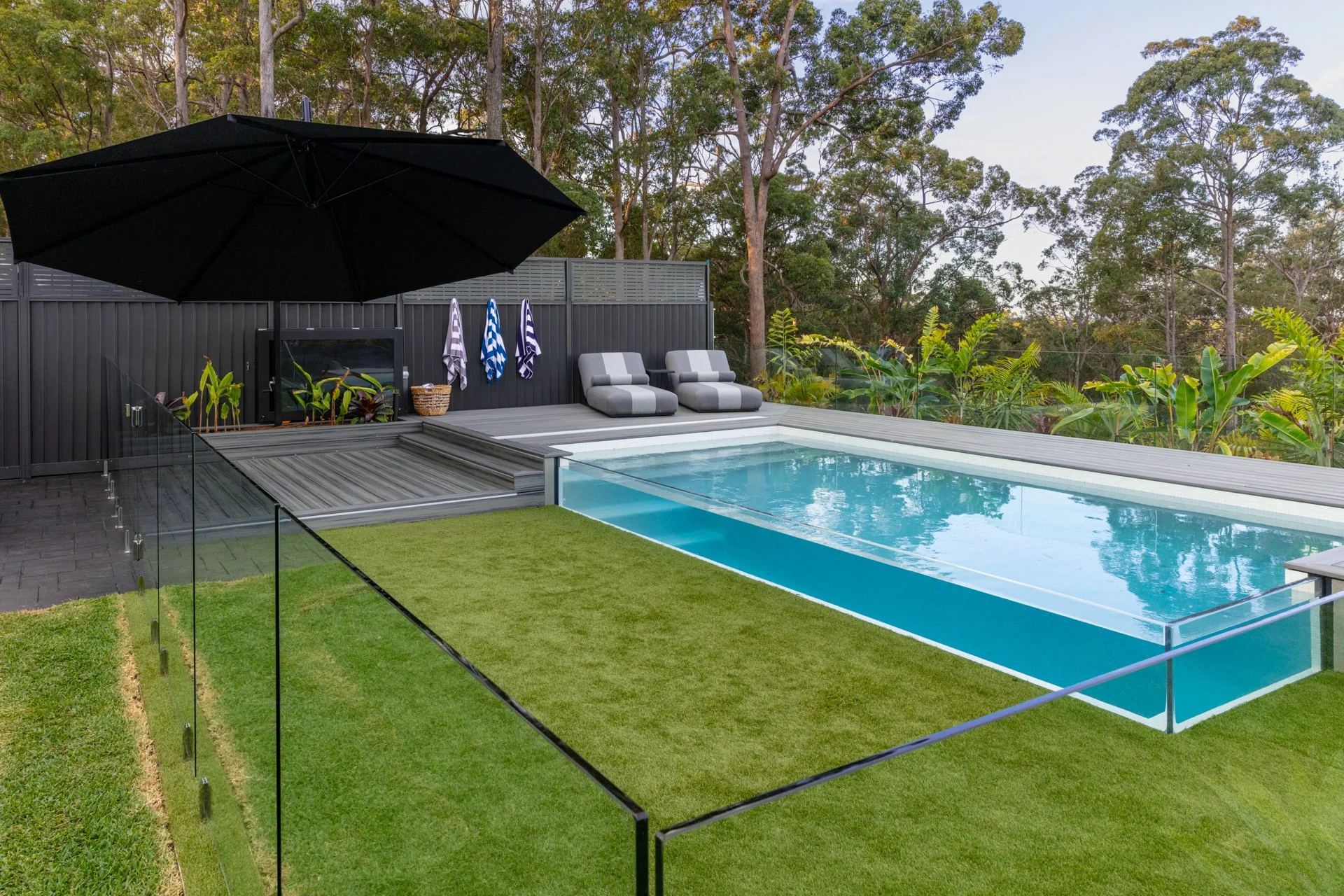 A backyard with a swimming pool, lounge chairs with striped cushions, a large black umbrella, and a glass fence, surrounded by tall trees and greenery.