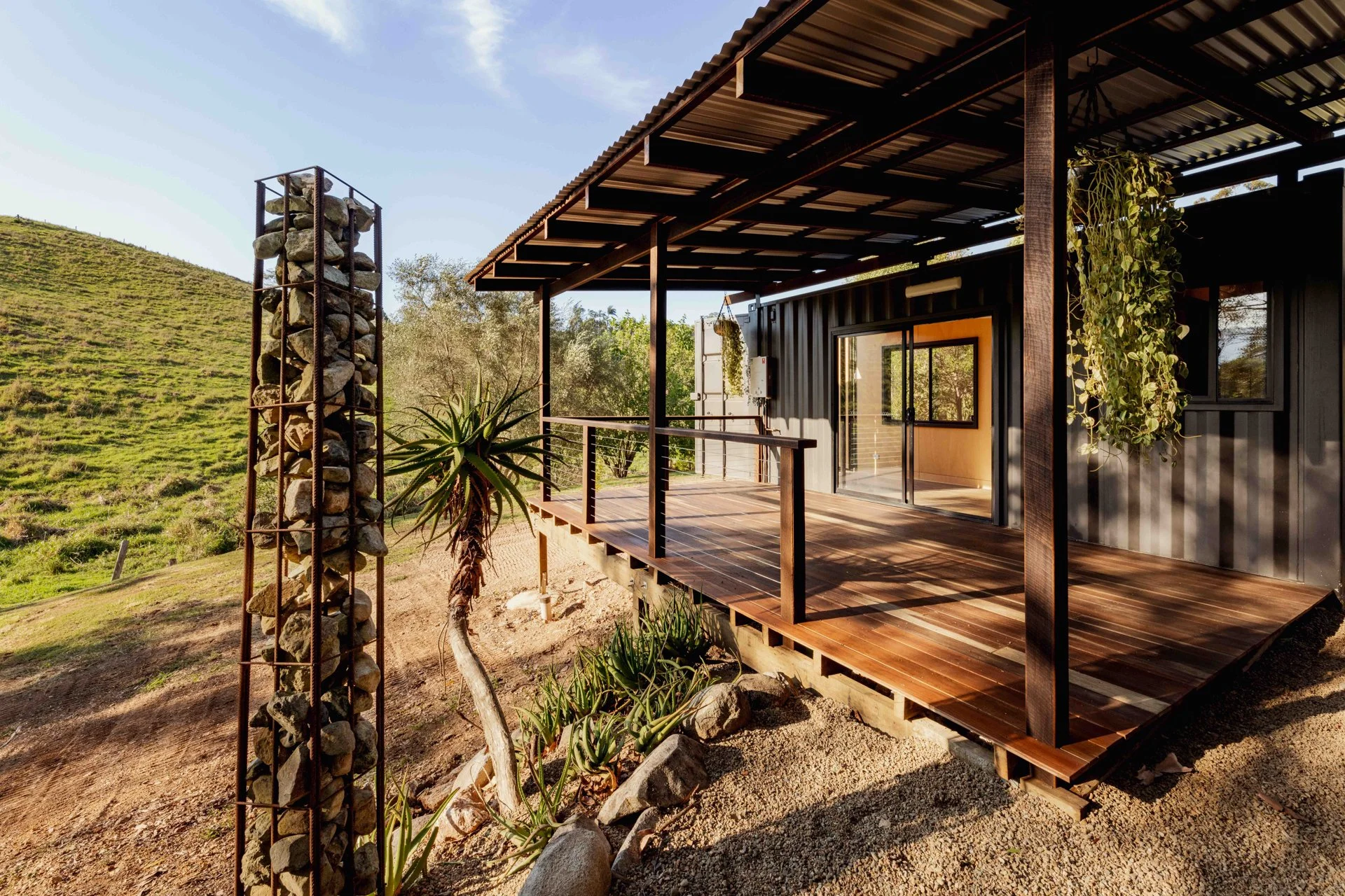 Modern house with a wooden deck and black exterior, large sliding glass doors, hanging plants, and outdoor stone and gardening features, set in a hilly green landscape under a clear blue sky.