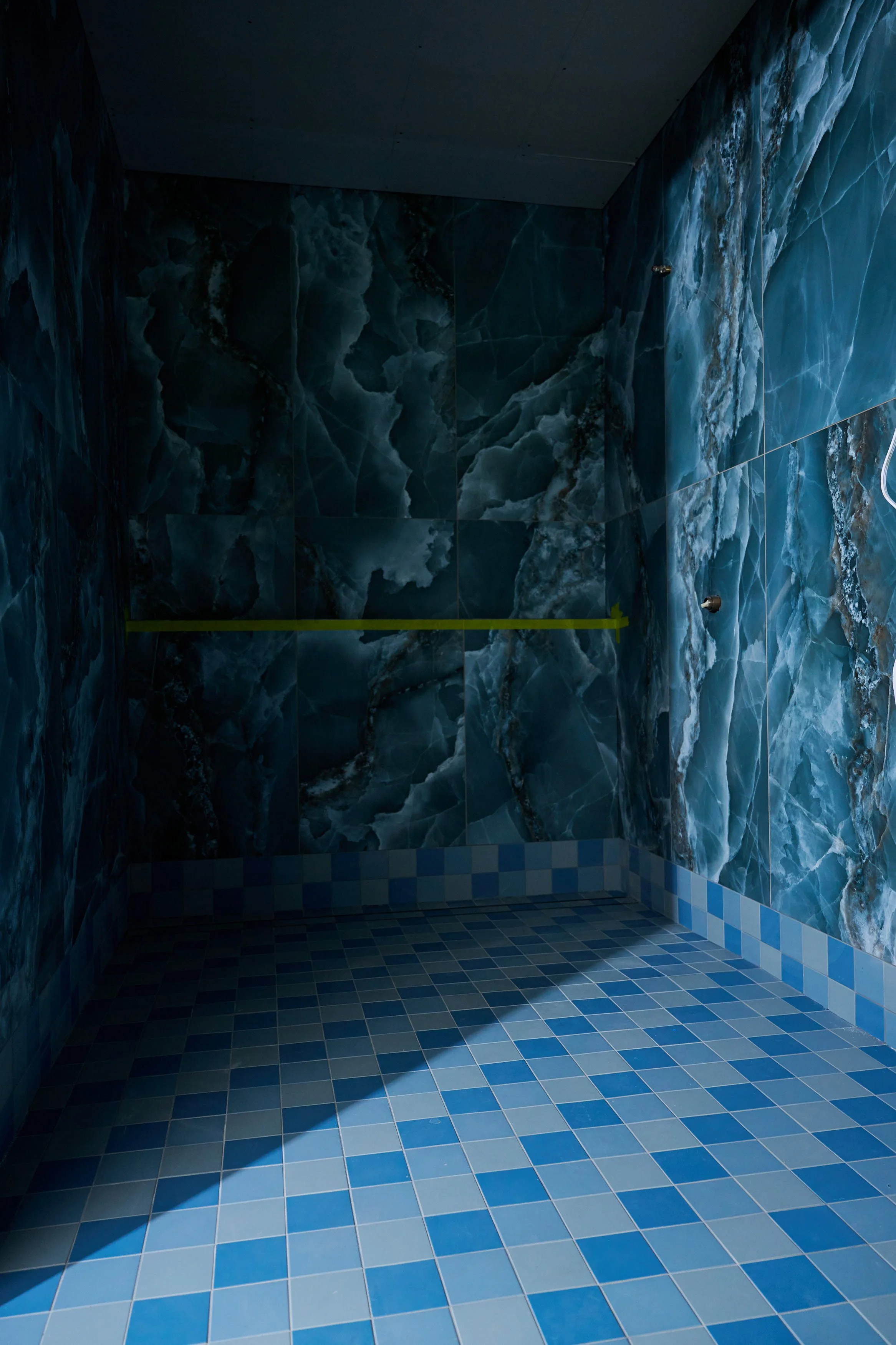 A tiled room with blue marble walls and blue checkered floor, with a yellow line across the wall and two shower head fixtures on the right wall.