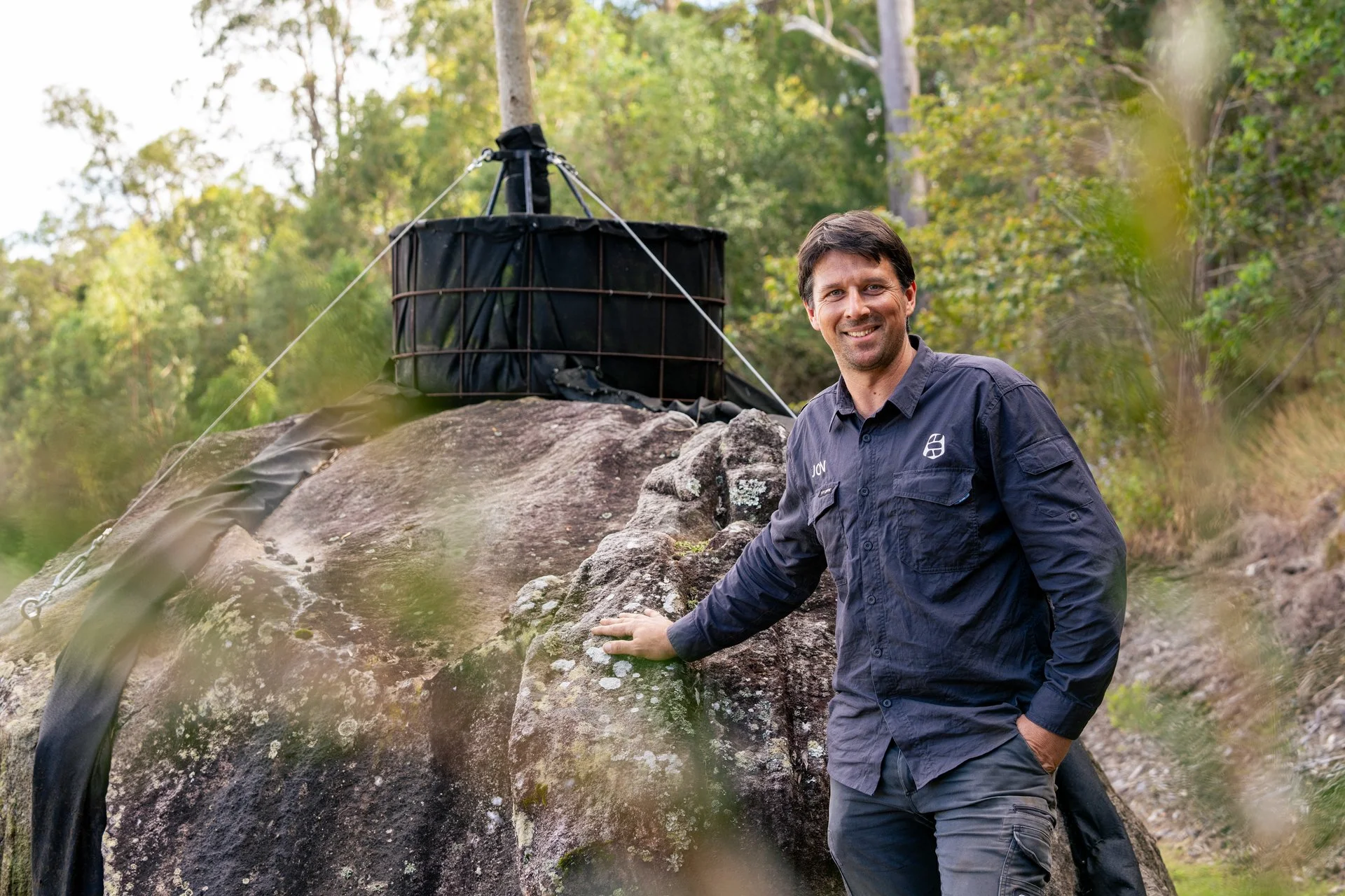 A man in a navy blue shirt with a logo, smiling outdoors in a forested area, standing beside a large rock with a black structure and water flowing over the edge.
