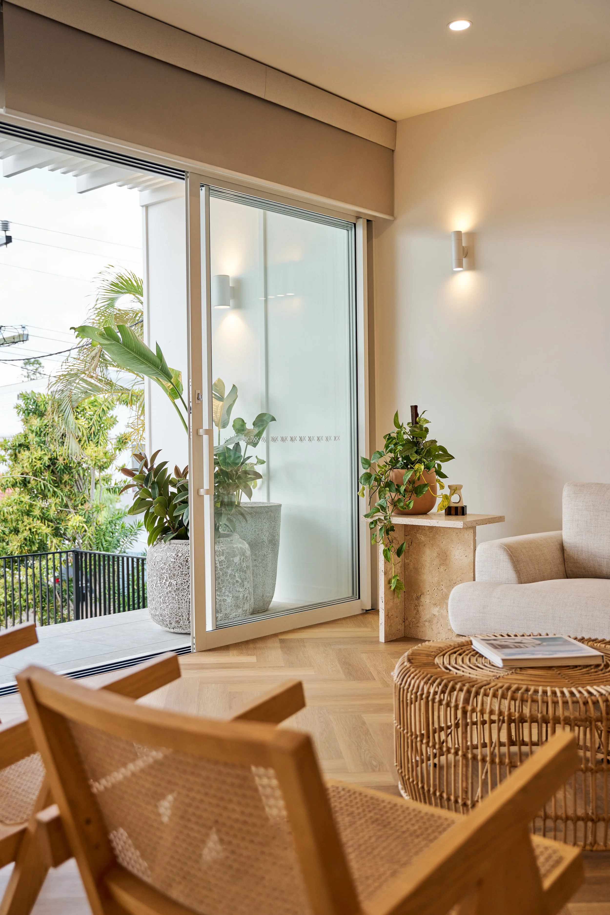 Living room with a sliding glass door open to a balcony, potted plants, a beige armchair, a wooden chair, and a rattan coffee table with magazines.