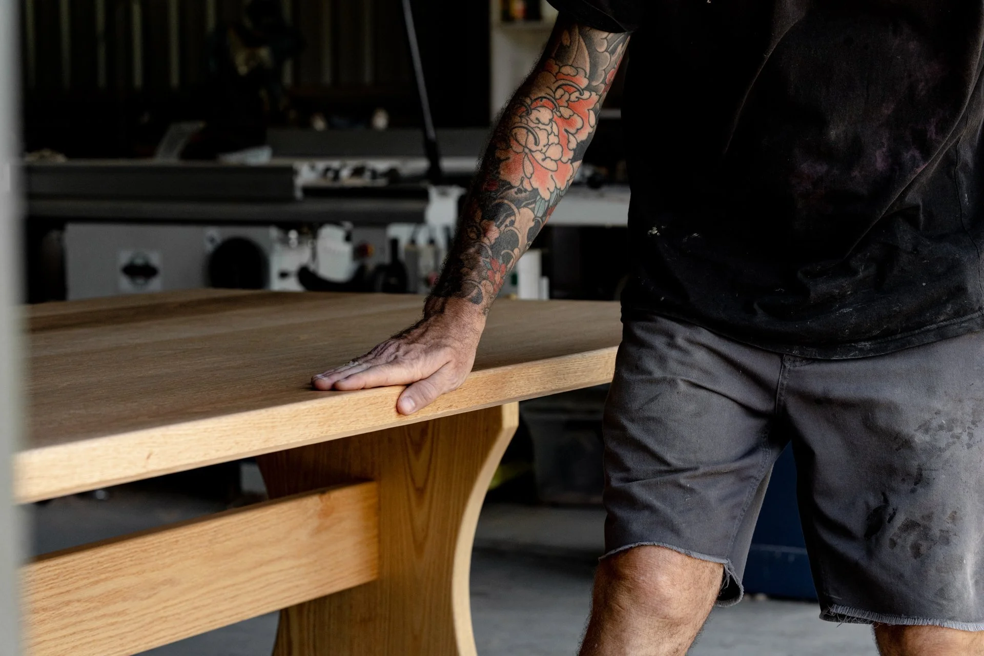 A man with a tattooed arm resting his hand on a wooden table in a workshop or studio setting.