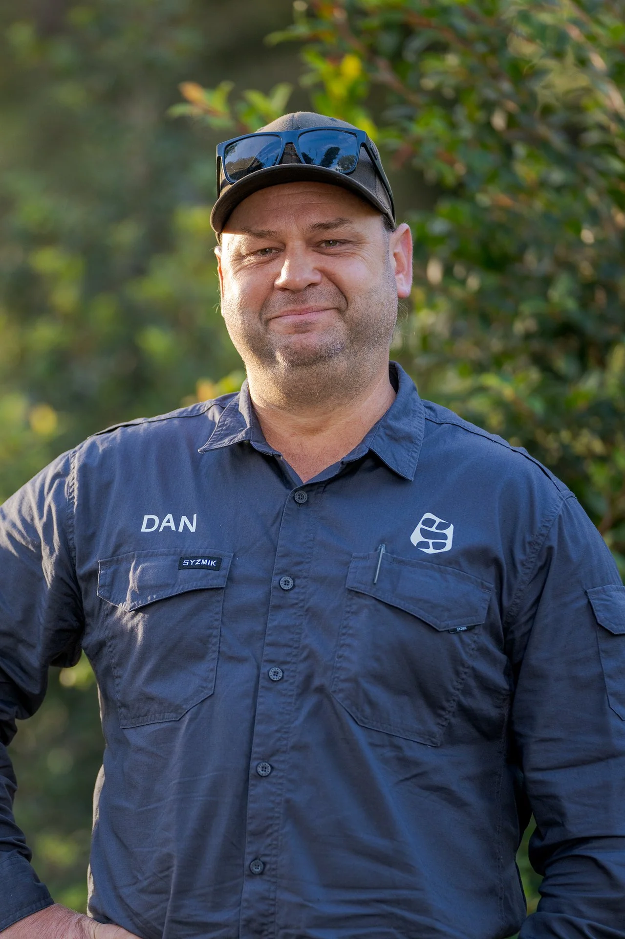 A man wearing a navy blue uniform with the name 'DAN' on it, sunglasses on his hat, and a control badge, standing outdoors with greenery in the background.