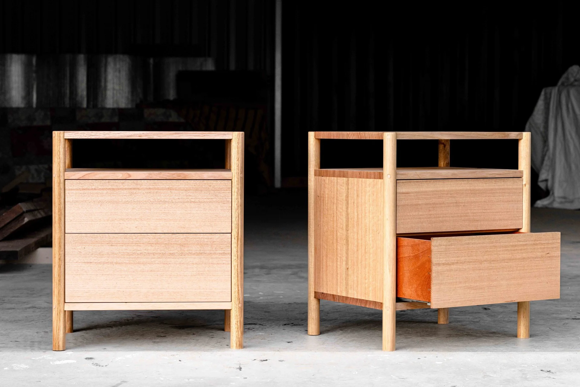 Two unfinished wooden nightstands with a drawer each, placed on a concrete floor in a workshop or storage area.