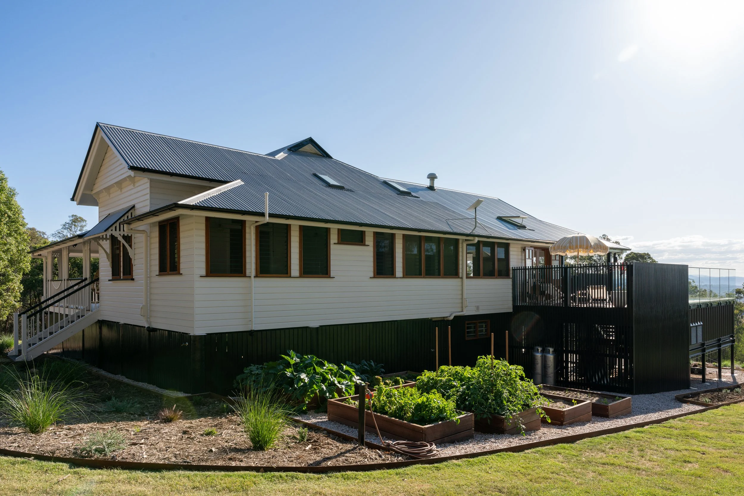 A two-story house with a metal roof, large windows, and a porch with stairs. There is a garden with raised beds and plants in front, and a fenced area to the right. The sky is clear and sunny.
