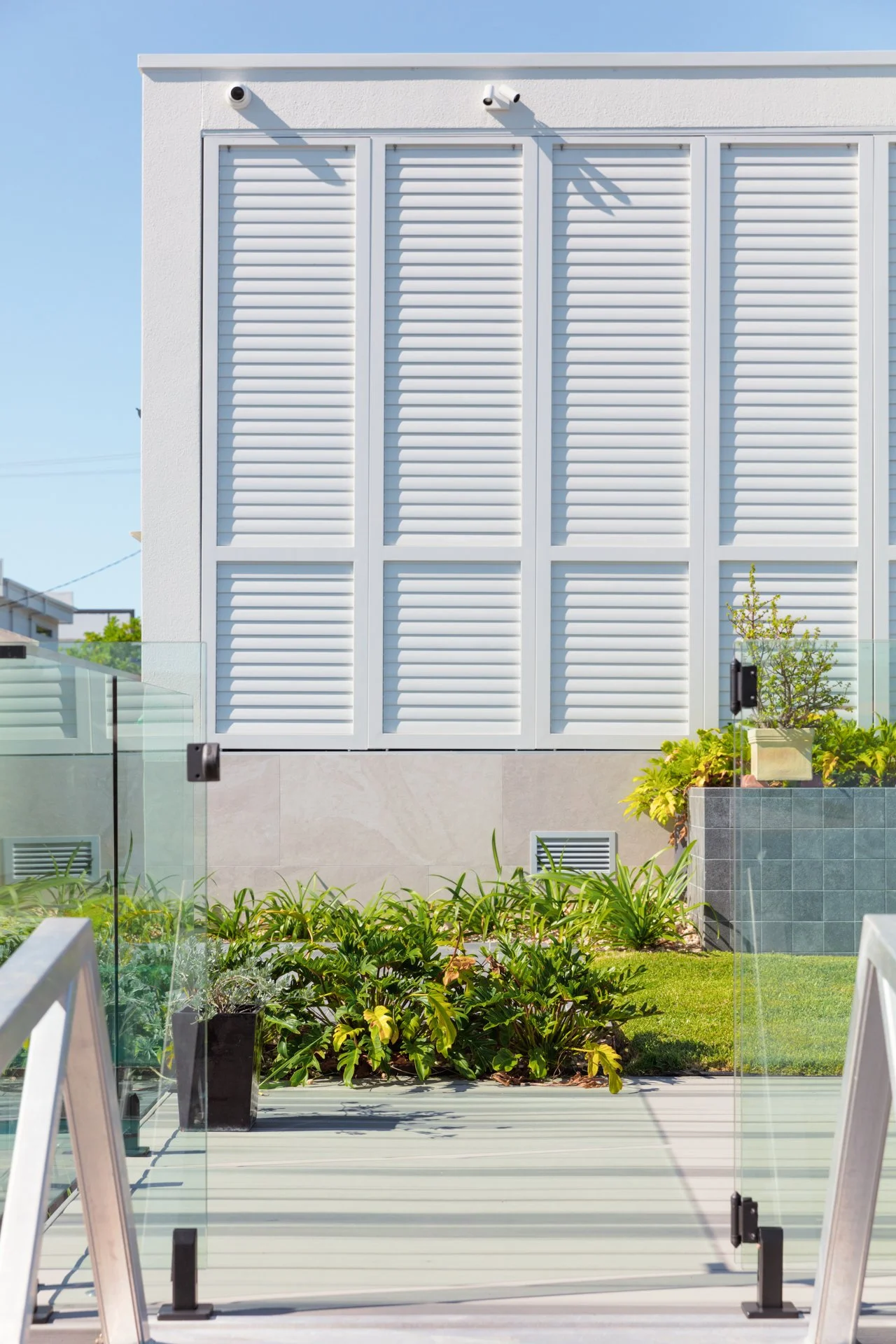 Modern building with white louvered shutters, greenery, and a glass railing on a terrace with a blue sky background.