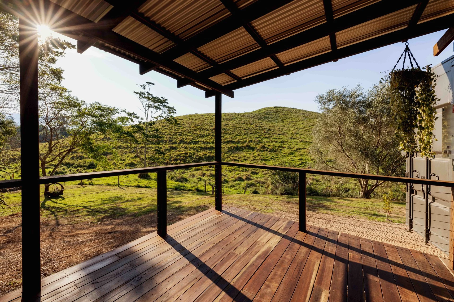 View from a porch with a wooden floor and black railing overlooking a grassy hillside with trees and a clear sky.