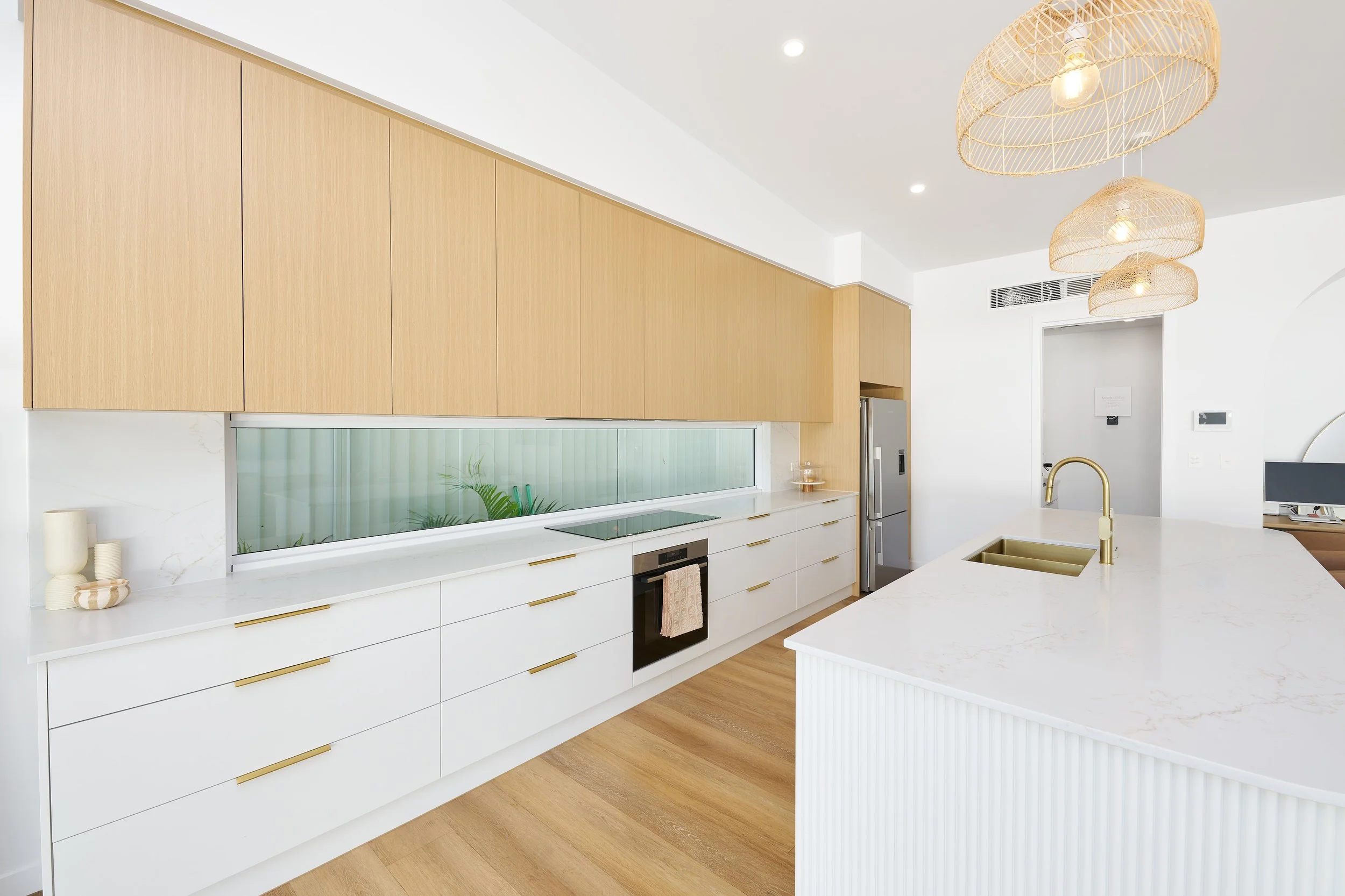 Modern kitchen with white and light wood cabinets, a marble countertop island with a gold faucet, pendant lights, and a refrigerator, featuring minimalist decor.