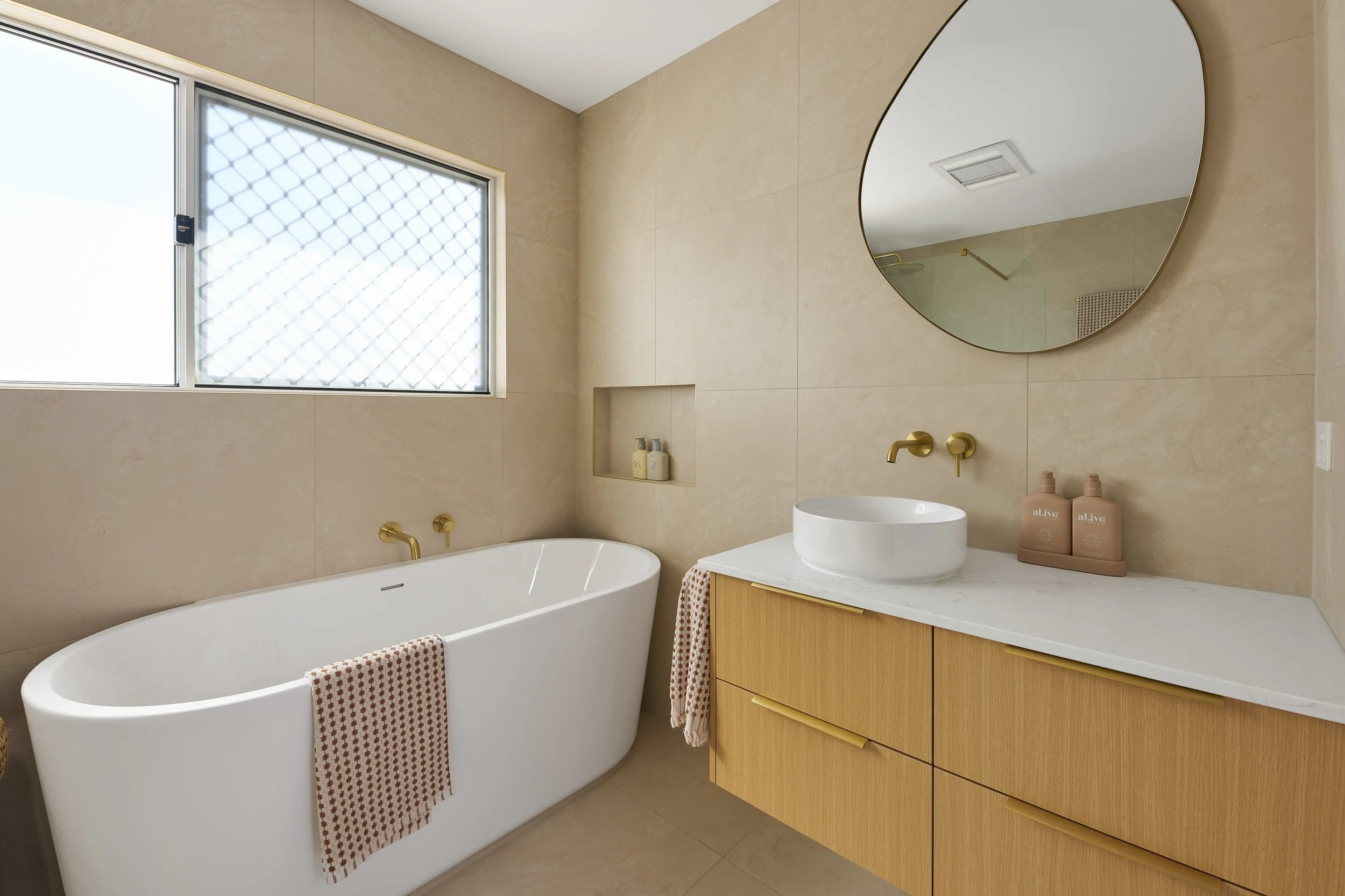 Modern bathroom with a freestanding bathtub, a wooden vanity with a white vessel sink, a round mirror, and a window with a security grate, all in neutral tones.