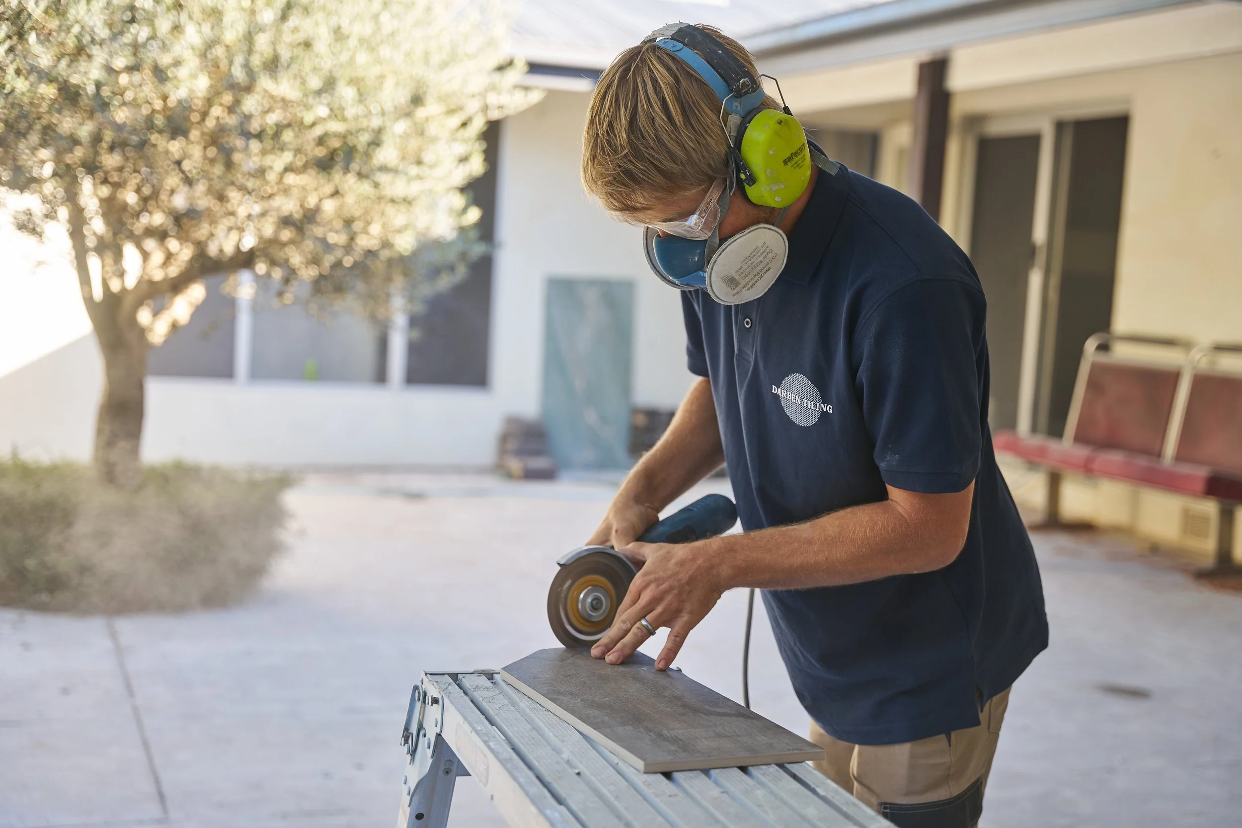 A man working outdoors at a construction site or workshop, using a power sander on a piece of wood. He is wearing safety ear protection, goggles, and a navy polo shirt with a logo that reads "DARBEN GUING." The background shows a tree, a building, a 