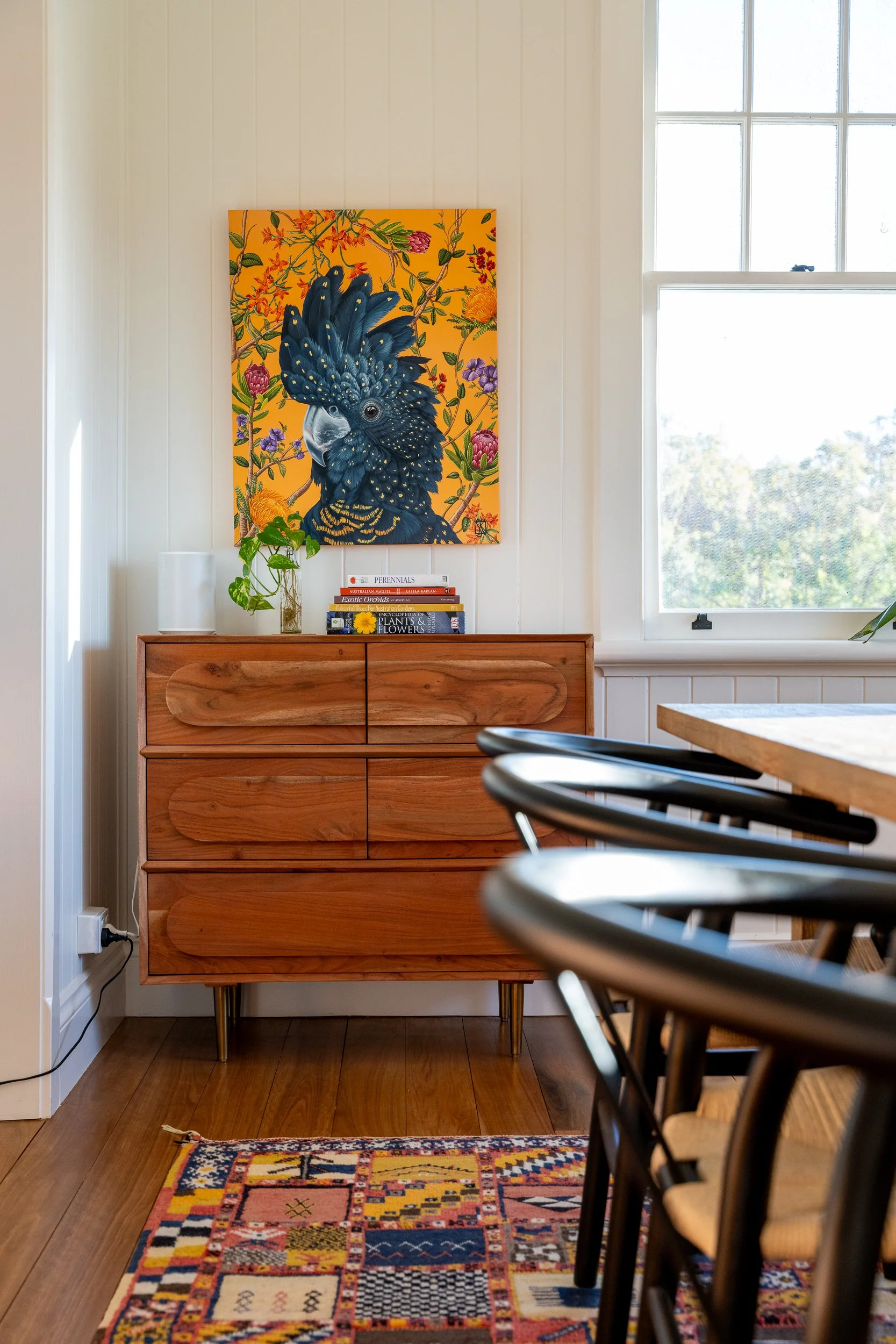 A wooden cabinet topped with a small potted plant, books, and a small picture frame, with a colorful patterned rug on the wooden floor in front of it, a large window letting in natural light, and a large painting of a black cockatoo with vibrant flor