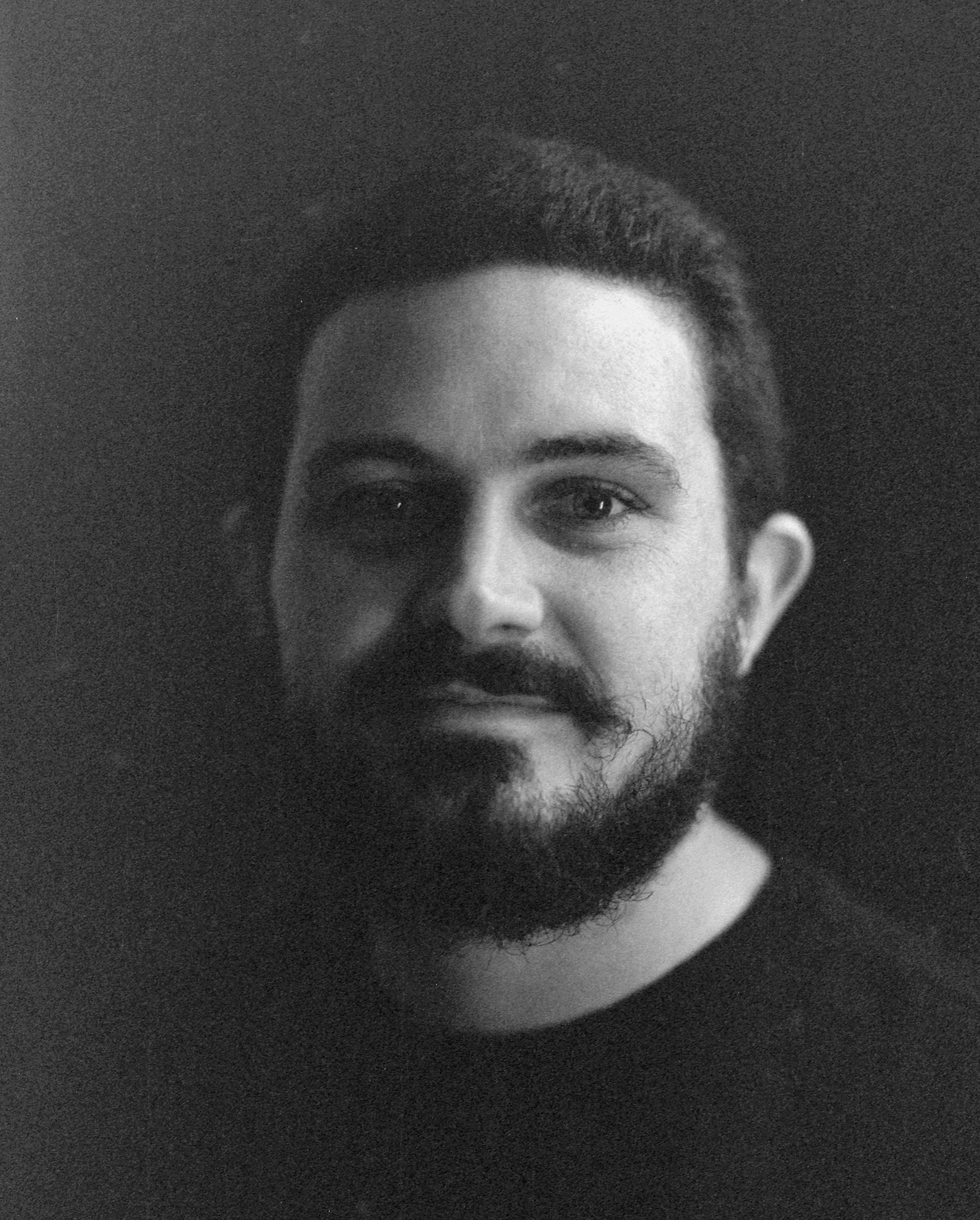 Close-up of the upper part of a young man's face, showing his eyes, nose, beard, and hairstyle, with a dark background.