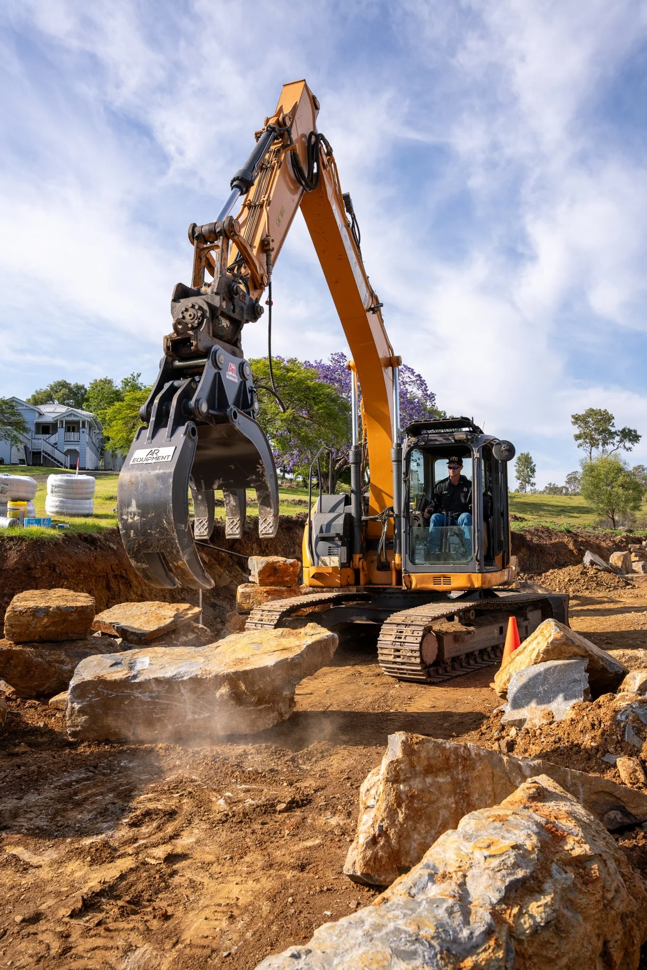 Construction excavator lifting a large rock on a construction site with dirt and rocks on the ground and residential houses in the background.