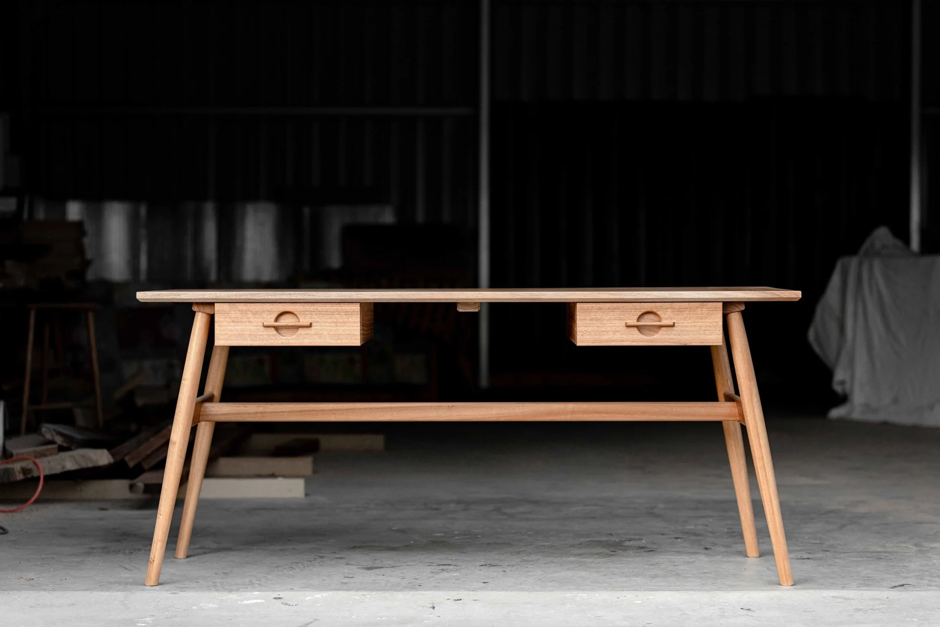 Wooden table with two small drawers in a workshop or garage with unfinished walls and various tools and furniture in the background.
