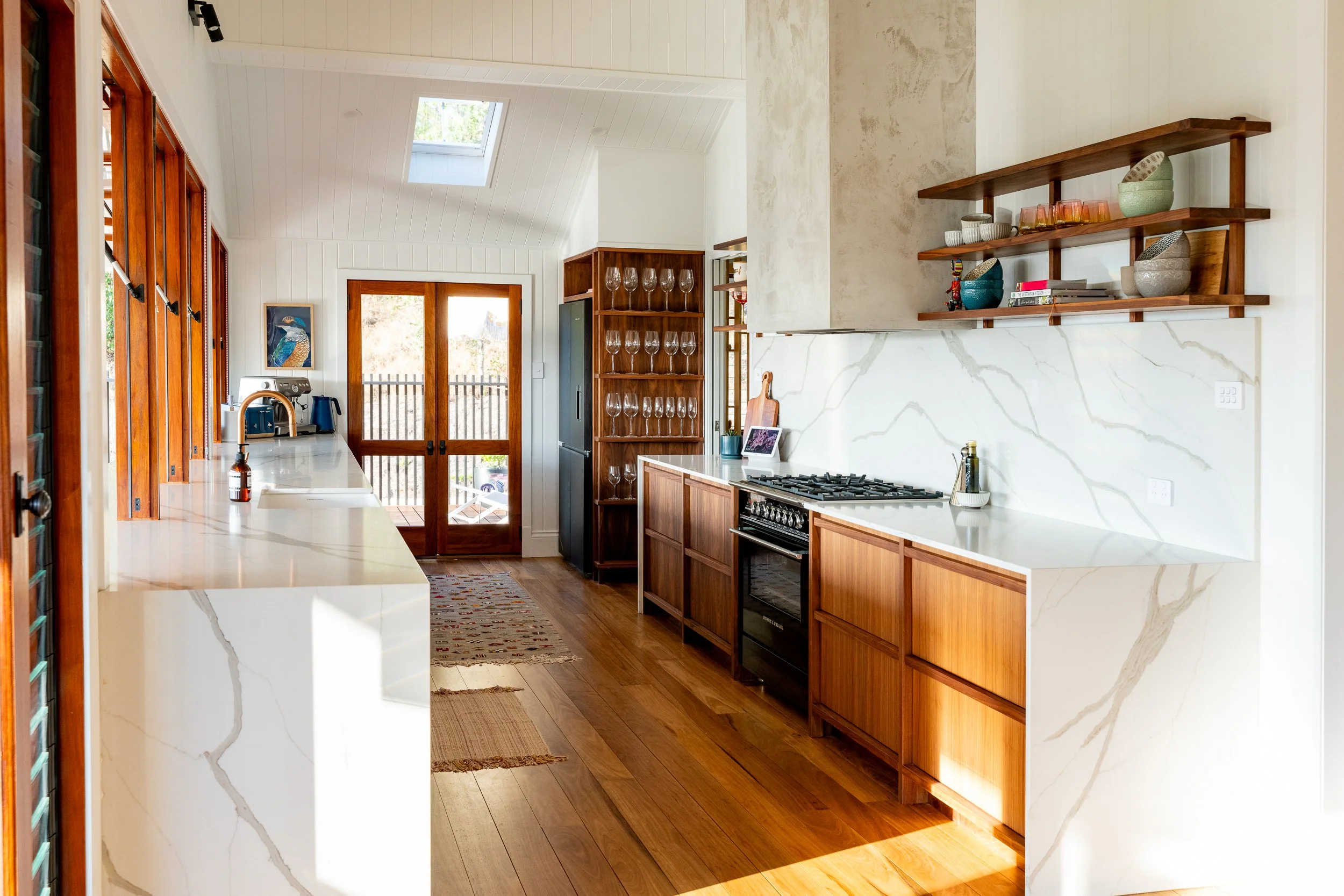 A modern kitchen with wooden cabinets, a white marble countertop, open wooden shelves holding dishes and bowls, a gas stove, and a gray refrigerator. There is a skylight in the ceiling letting in natural light, and a door leading outside with a view 