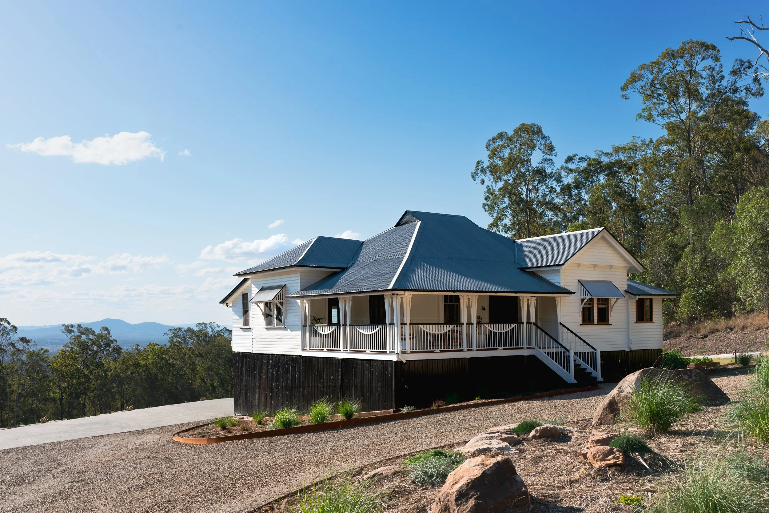 A white house with a black foundation and a covered front porch with white railings, situated on a hillside. There is a gravel driveway and some decorative rocks and plants in the foreground. Tall green trees and a mountain range can be seen in the b