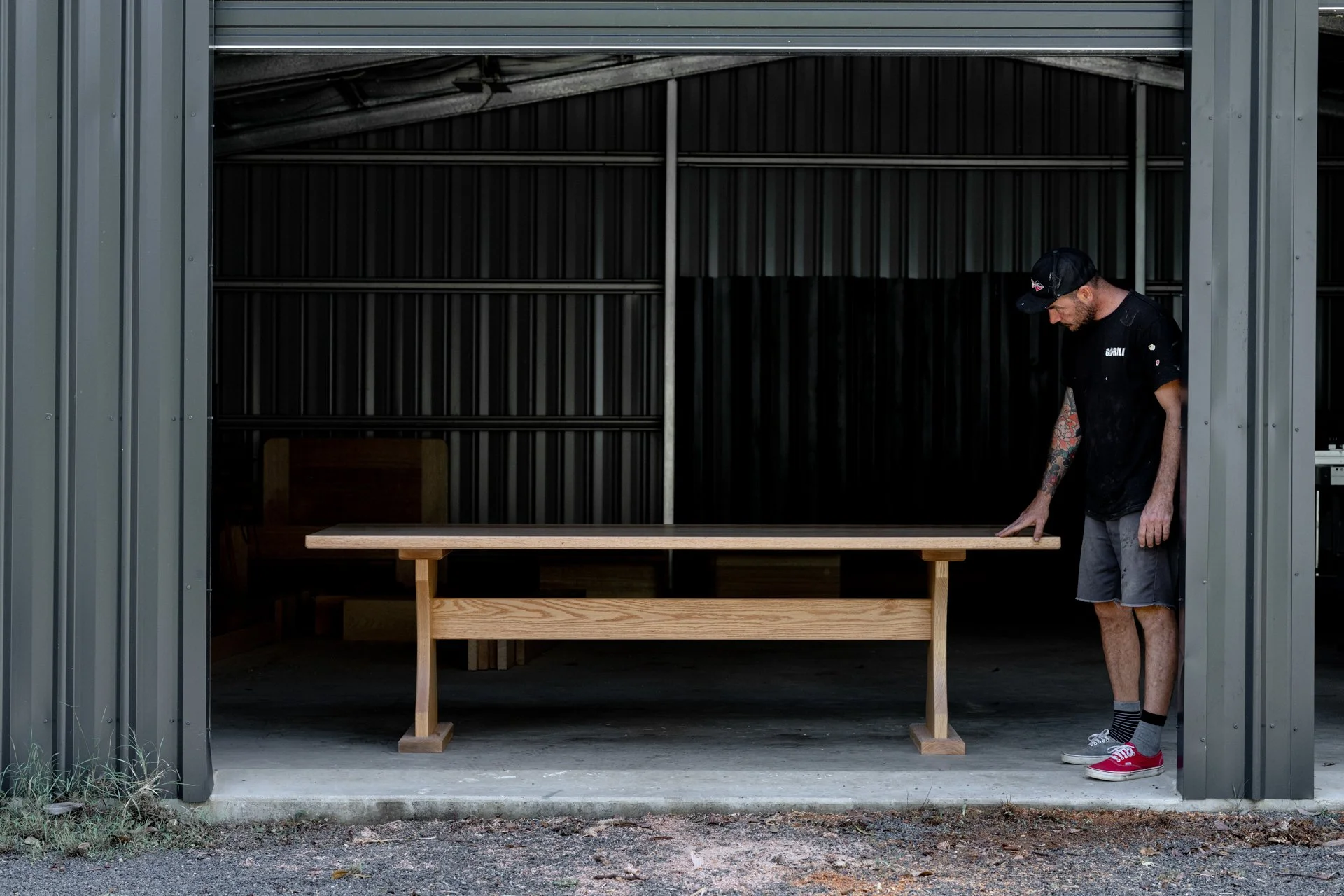 A man with tattoos, wearing a black cap, black t-shirt, and gray shorts, stands inside a metal storage shed, looking down at a wooden table in front of him, with the shed door open.