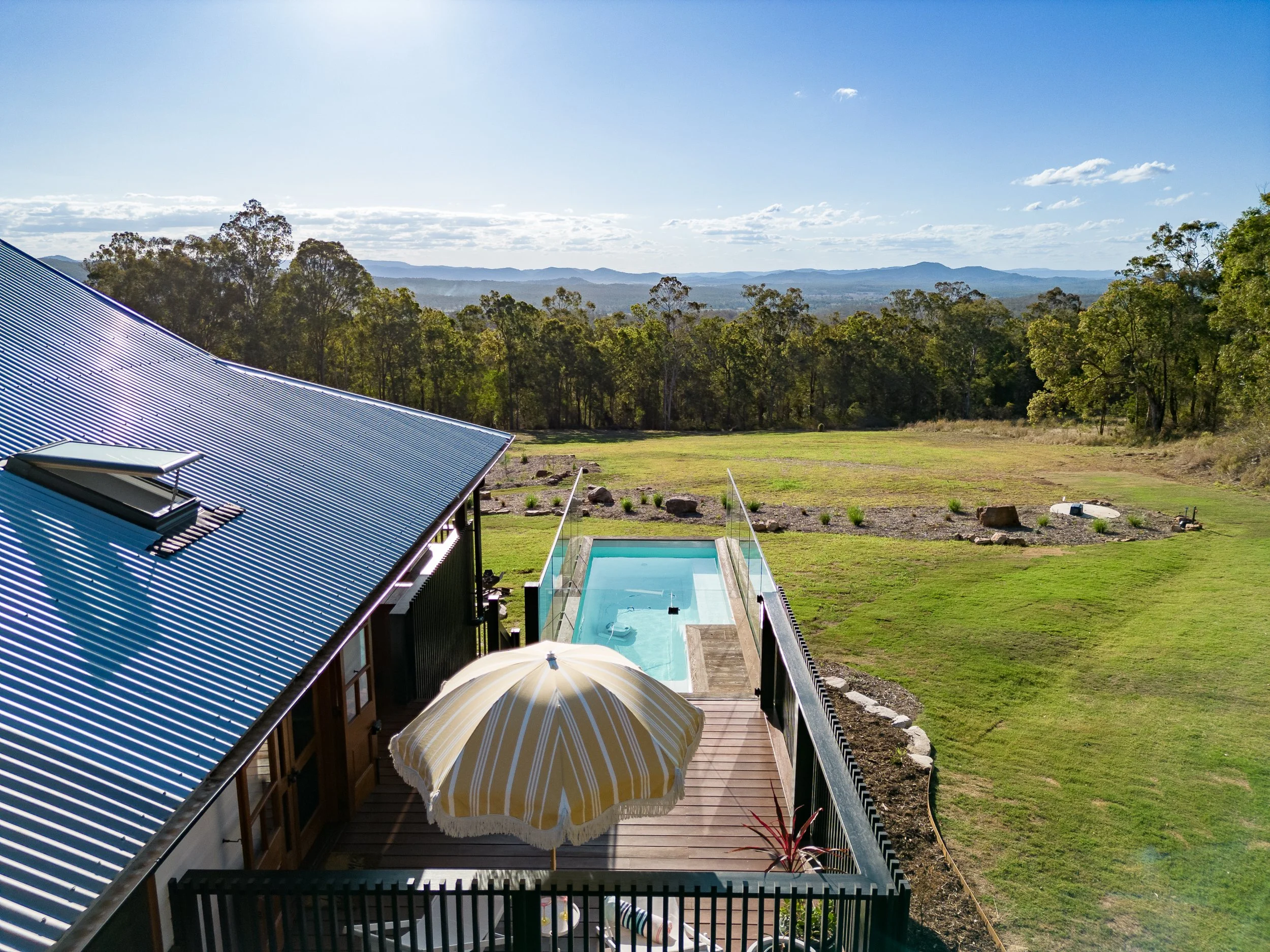 A view from a deck showing a swimming pool, a large parasol, a grassy yard, and a background of forest and distant mountains under a partly cloudy sky.