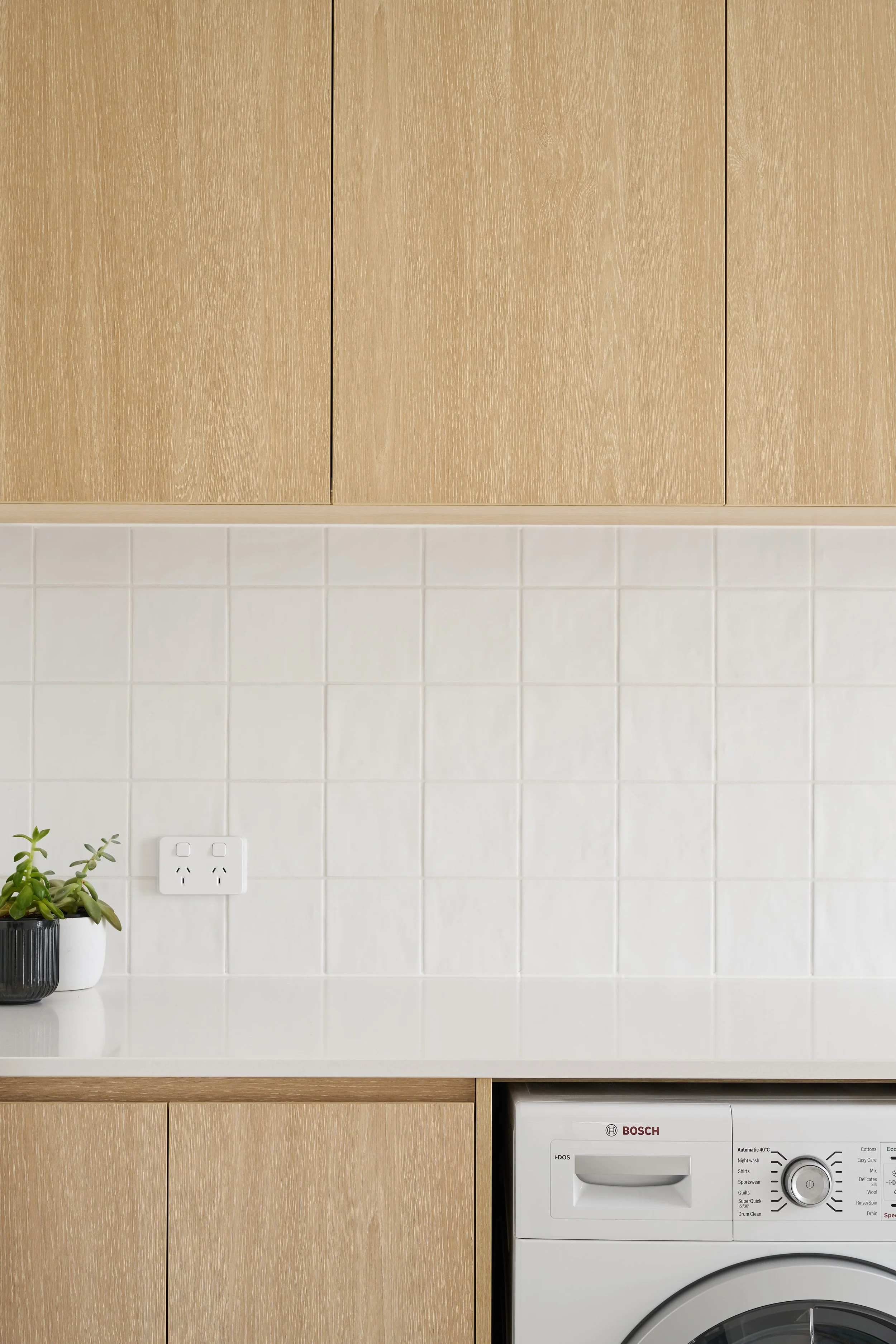 A kitchen scene featuring a white countertop with a small potted plant, a white electrical outlet, and a Bosch washing machine, with wooden cabinets above that have a light wood finish, and a white tiled backsplash.