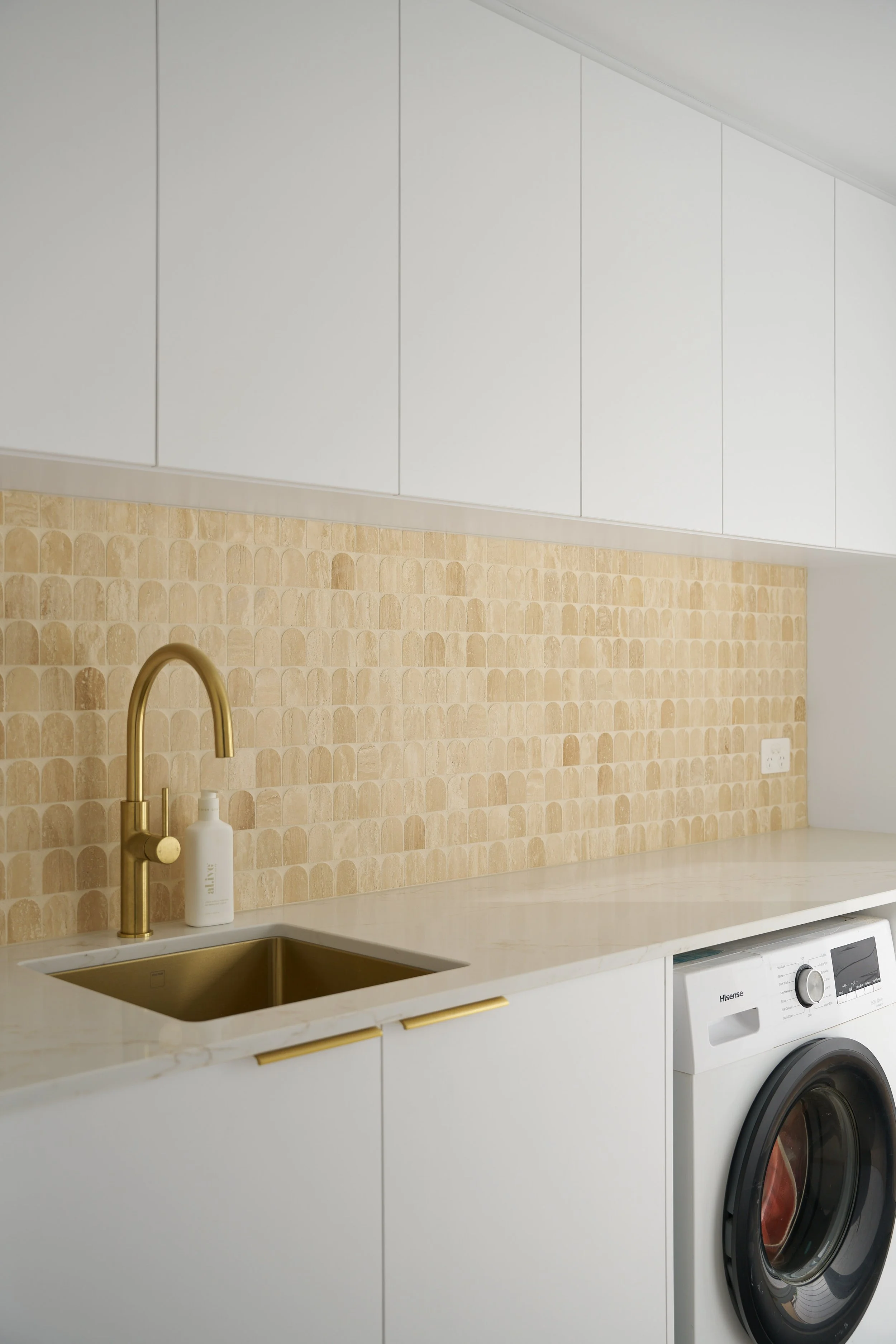 Modern laundry room with white cabinets, a beige tiled backsplash, a gold faucet, and a front-loading washing machine.
