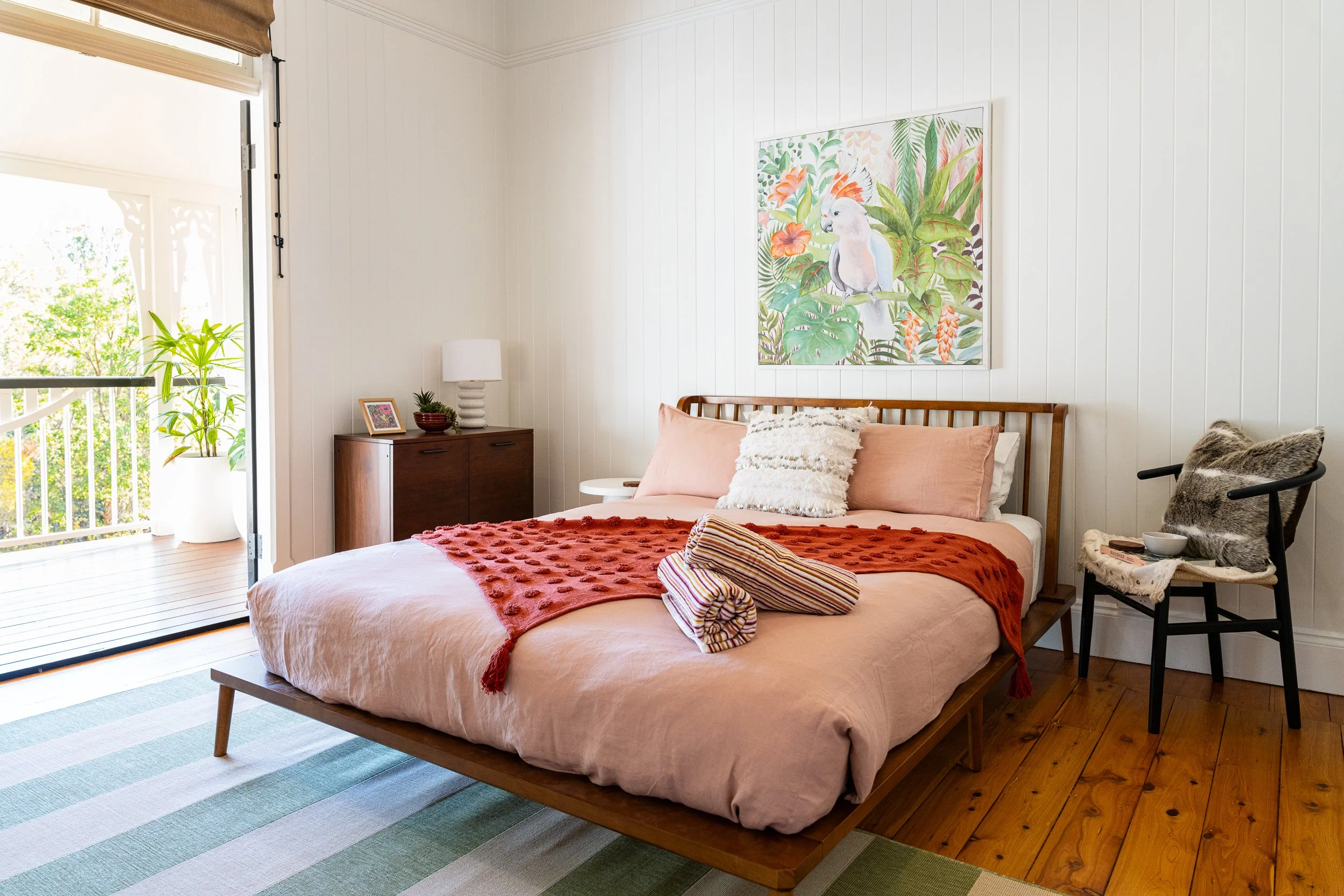 A bedroom with a wooden bed frame, peach bedding, and a burnt orange blanket. There are white and textured pillows on the bed. To the right, there's a chair with a furry blanket and a small table with bowls. A framed photo and a lamp sit on a wooden 