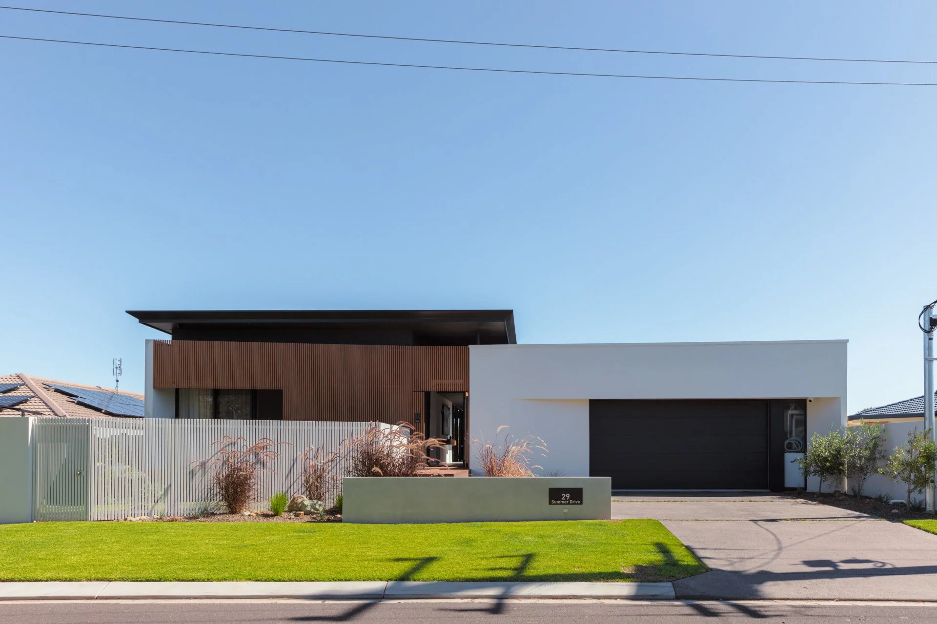 Modern house with a flat roof, a white and brown facade, and a black garage door, surrounded by a green lawn and a white fence under a clear blue sky.