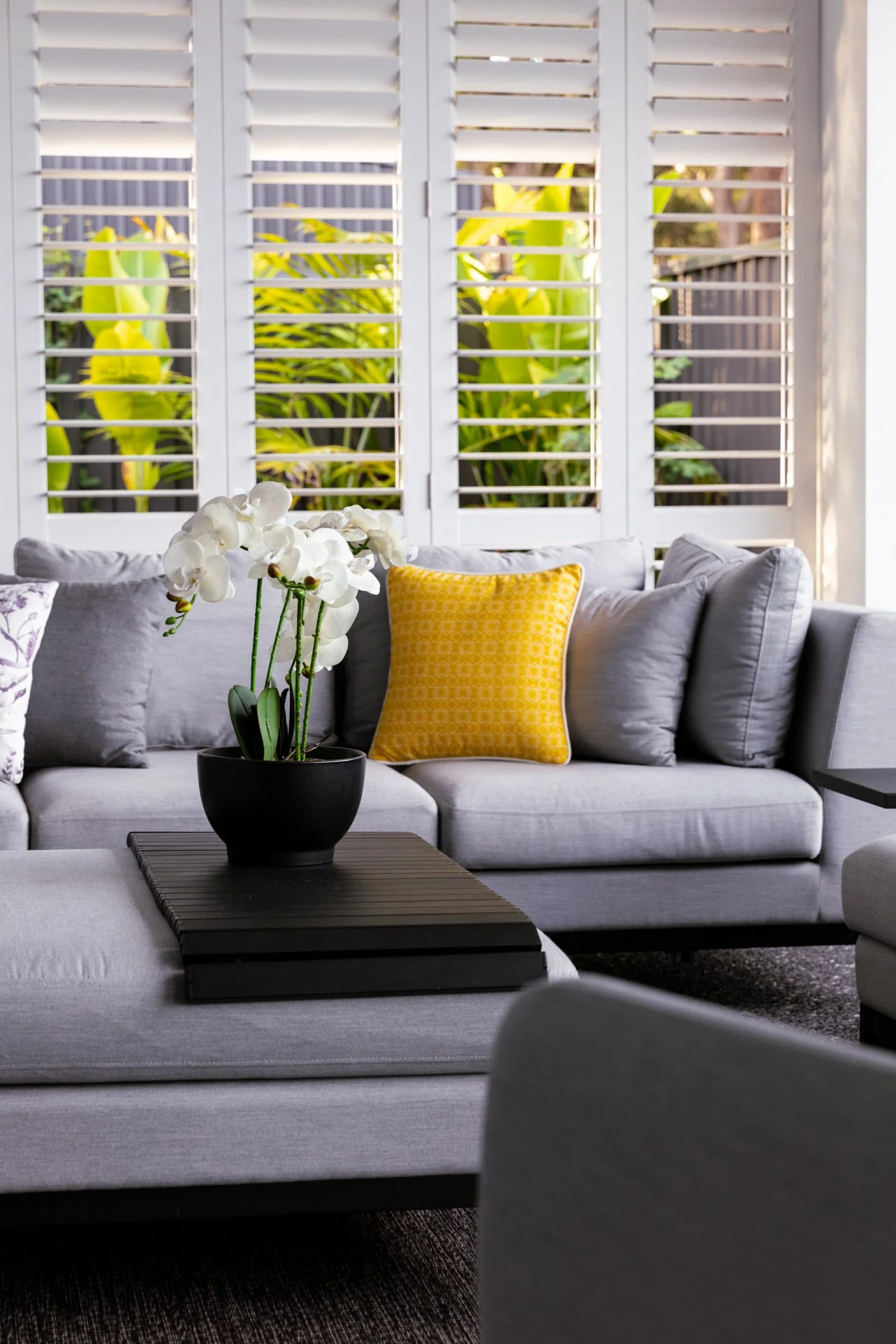 Living room with white shutters, a light gray sofa with a yellow pillow, a black coffee table with a potted white orchid, and a window view of green plants outside.