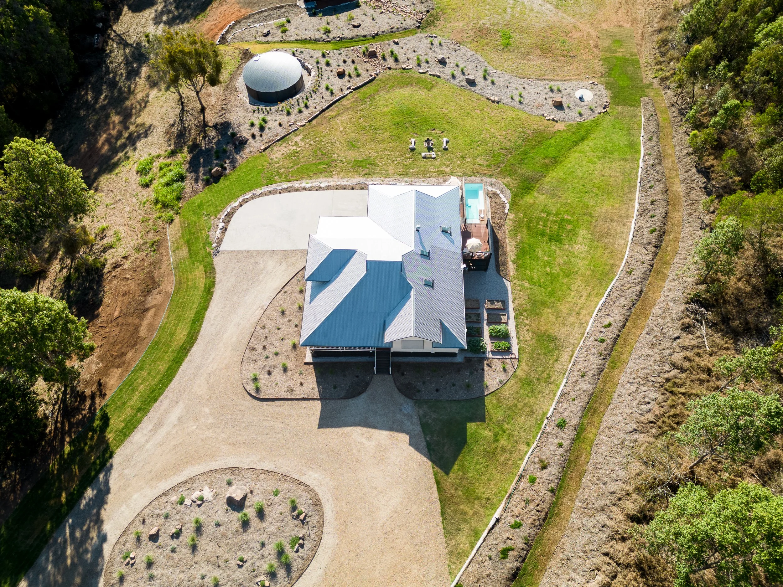 An aerial view of a house with a grey roof, a backyard with a swimming pool, a concrete patio, a lawn, and landscaped garden beds. There is a curved gravel driveway leading to the house, surrounded by trees and a fence.