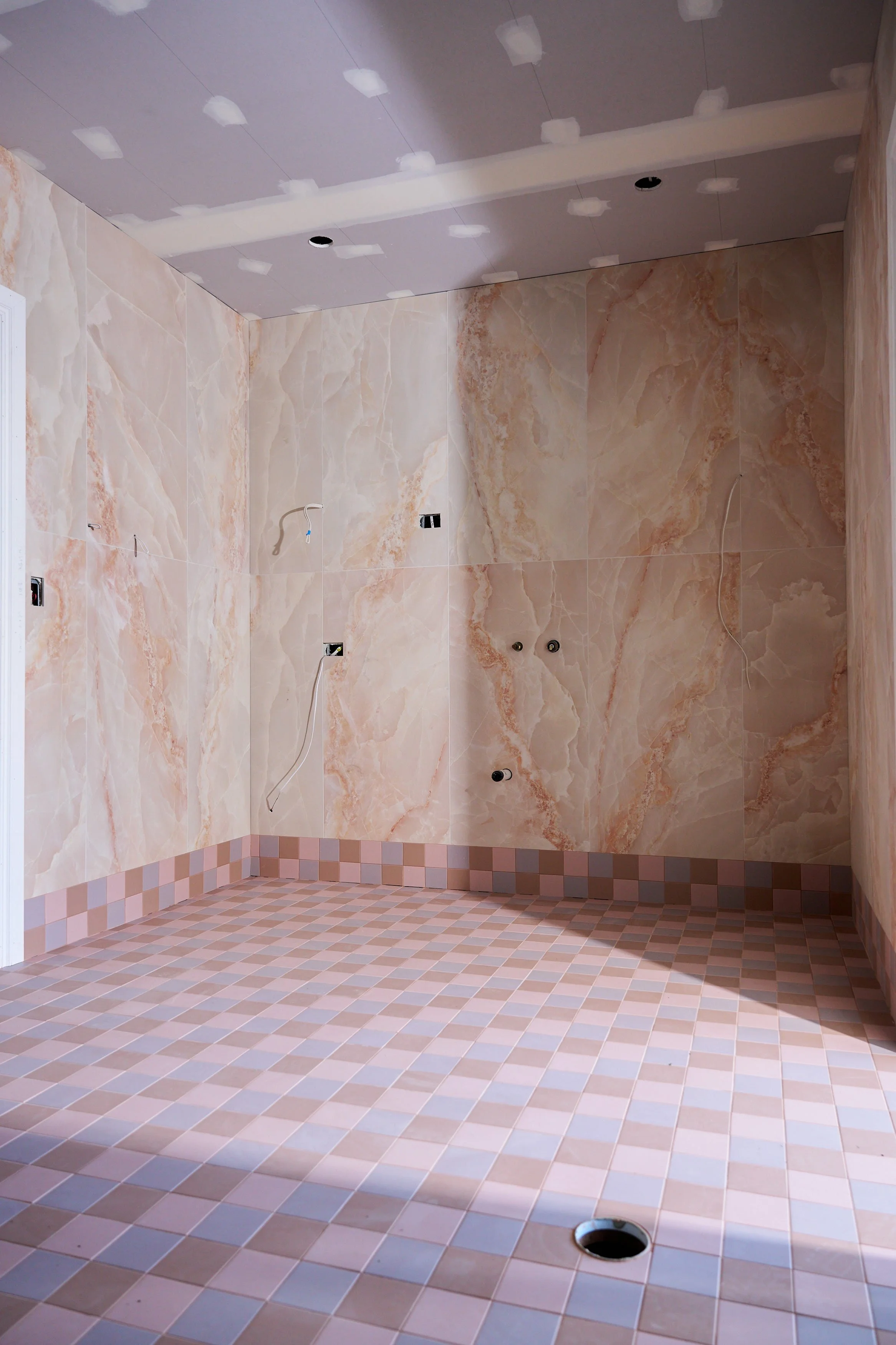 Empty shower area with beige marble wall tiles and a pink and beige checkered floor with a round drain in the corner.