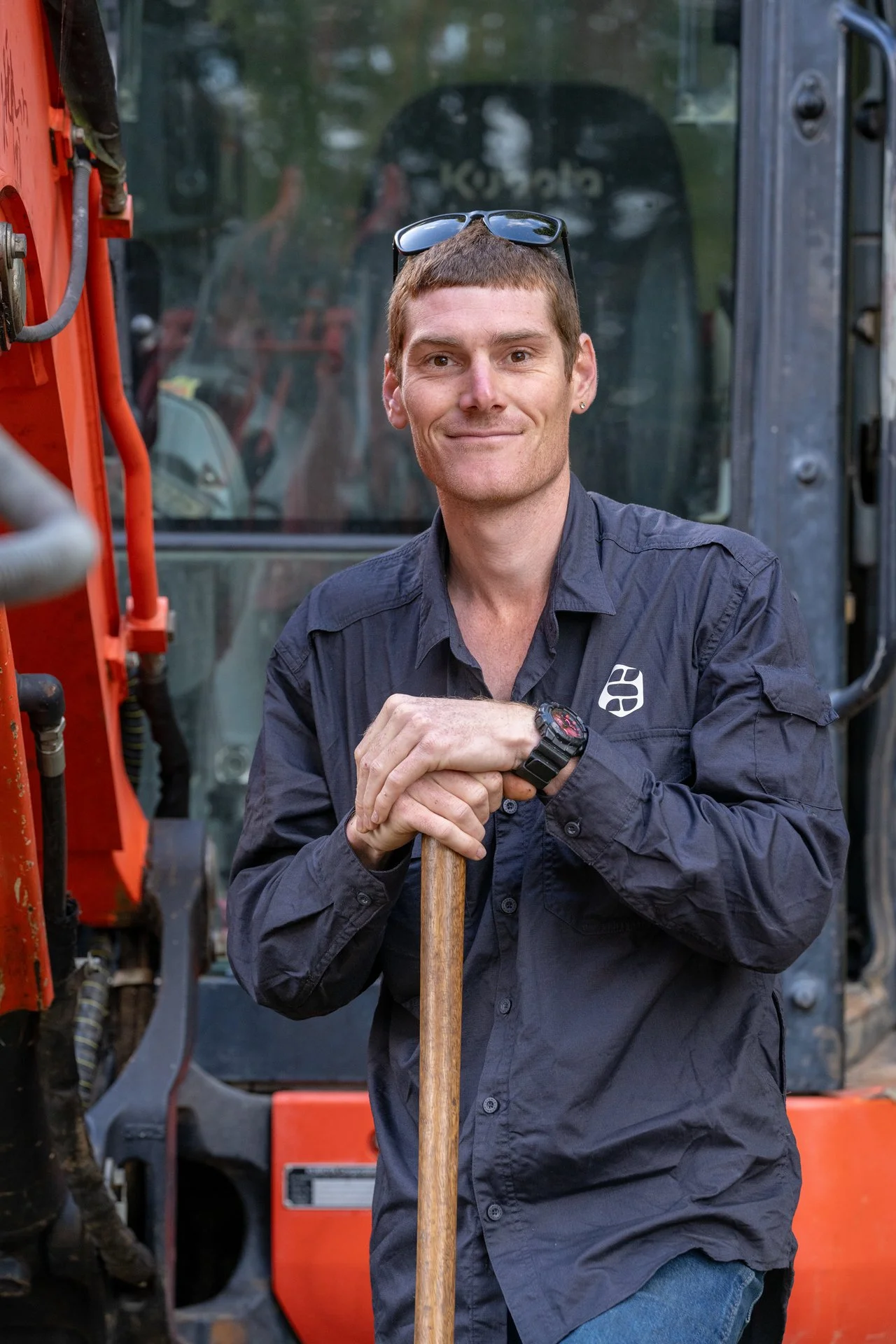 A man with short brown hair and sunglasses on his head, wearing a black long-sleeve shirt with a logo, posing outdoors in front of a construction vehicle, holding a shovel.