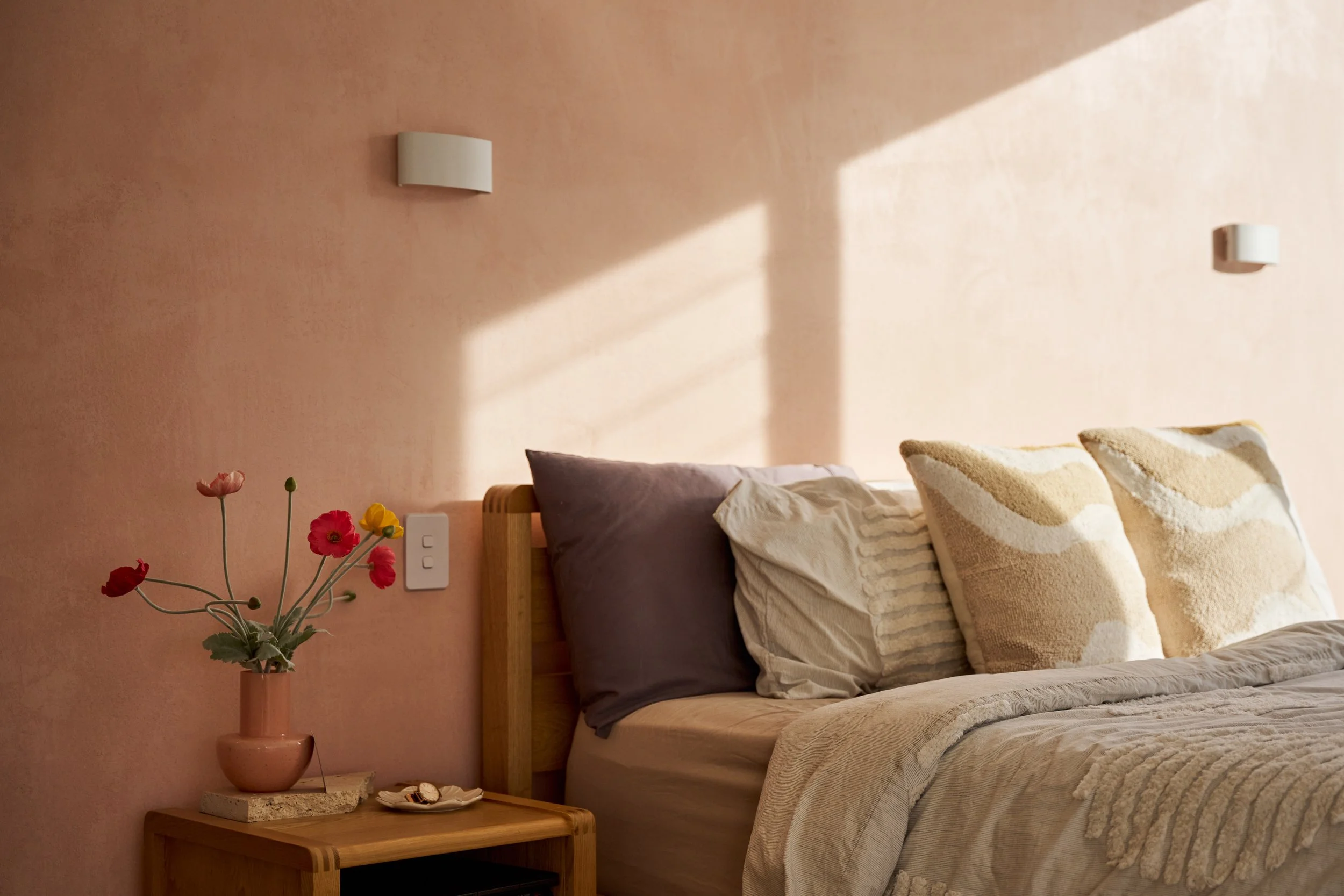 Bedroom with pink walls, a wooden bed with beige and purple pillows, a wooden side table with a pink vase holding red, pink, and yellow flowers, and a wall-mounted white light fixture.