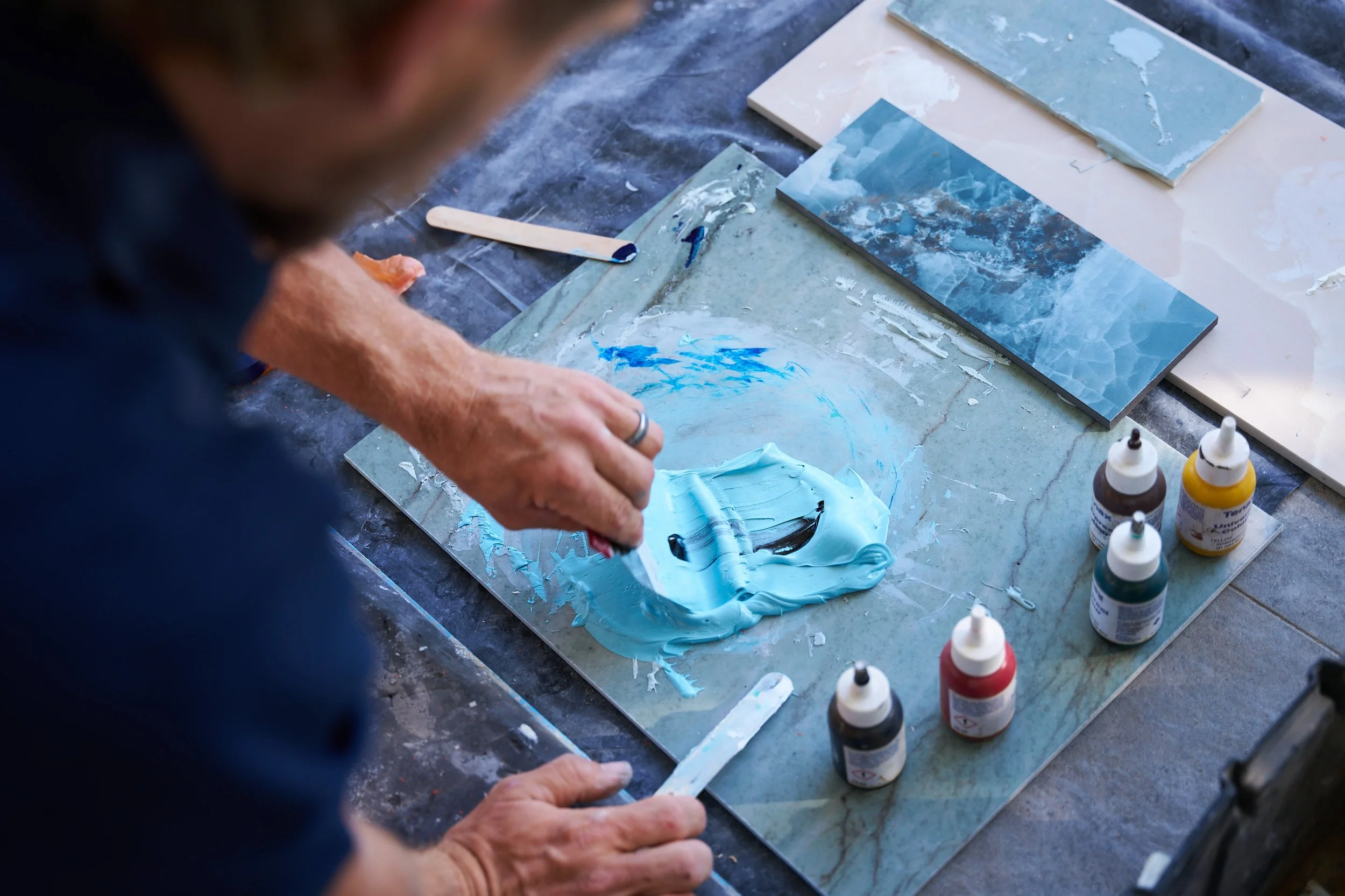 Person working on a craft project with tiles and paint, applying blue paint to a tile surface.