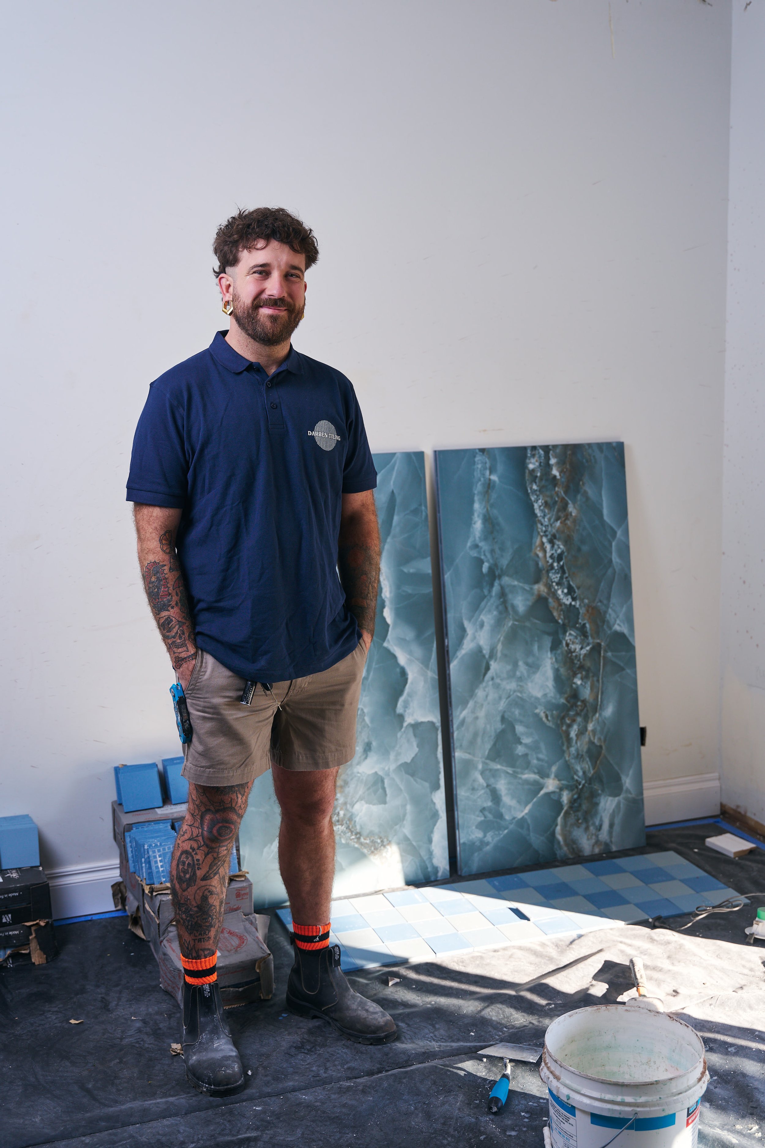 A man with tattoos on his arms and legs, wearing a blue polo shirt, beige shorts, and black boots and orange and black striped socks, standing in a room with two large blue marble-patterned tiles behind him. The room appears to be under construction 