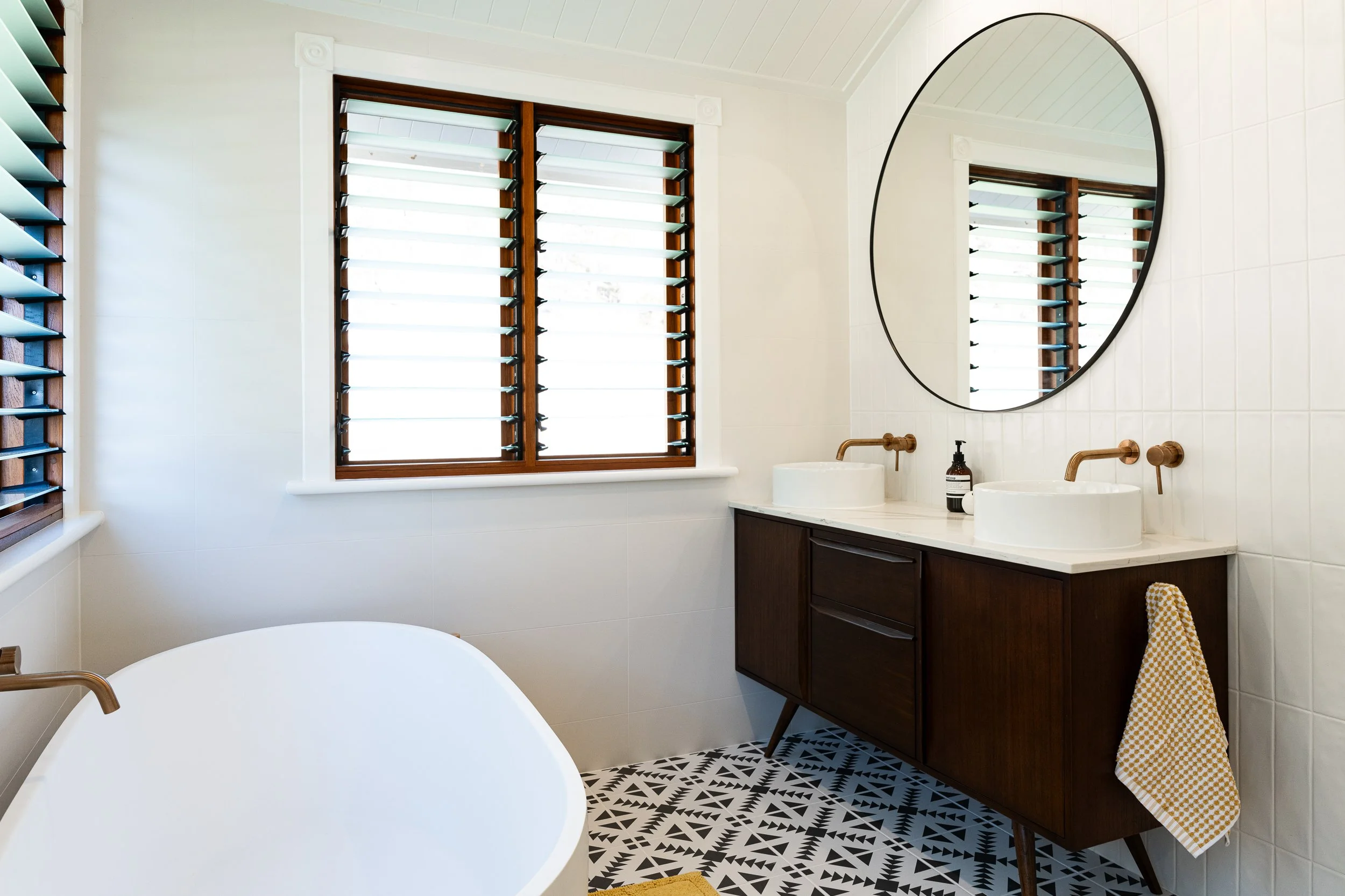 A bathroom with a round mirror above a double sink vanity, wooden cabinetry, a soap dispenser, plaid yellow and white towel, patterned floor tiles, a window with wooden shutters, and a corner shelf with teal boxes.