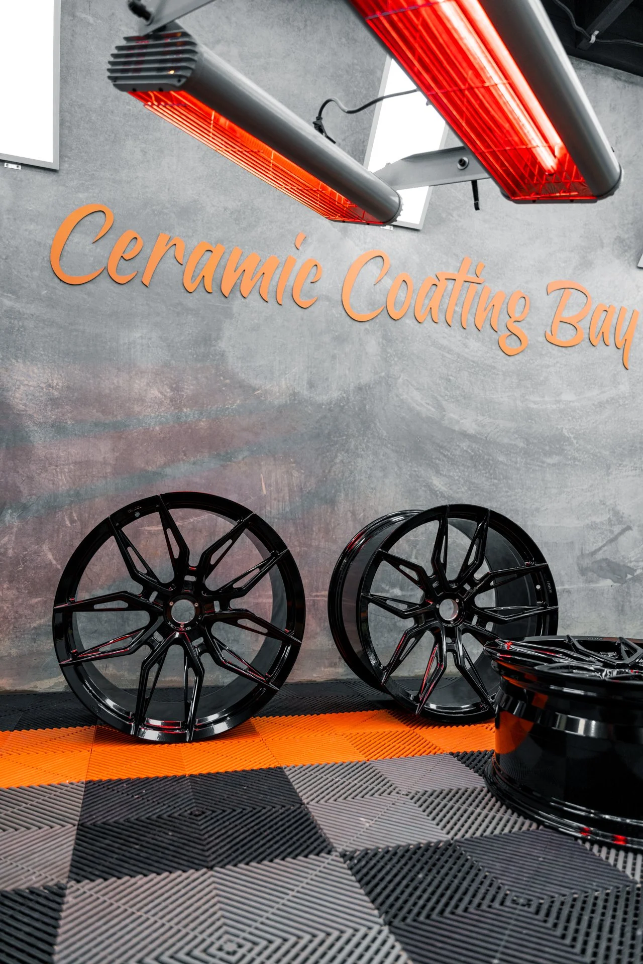 A garage or workshop with black car rims on a patterned gray and black checkered floor, and overhead infrared heat lamps. A sign on the wall reads "Ceramic Coating Bay."