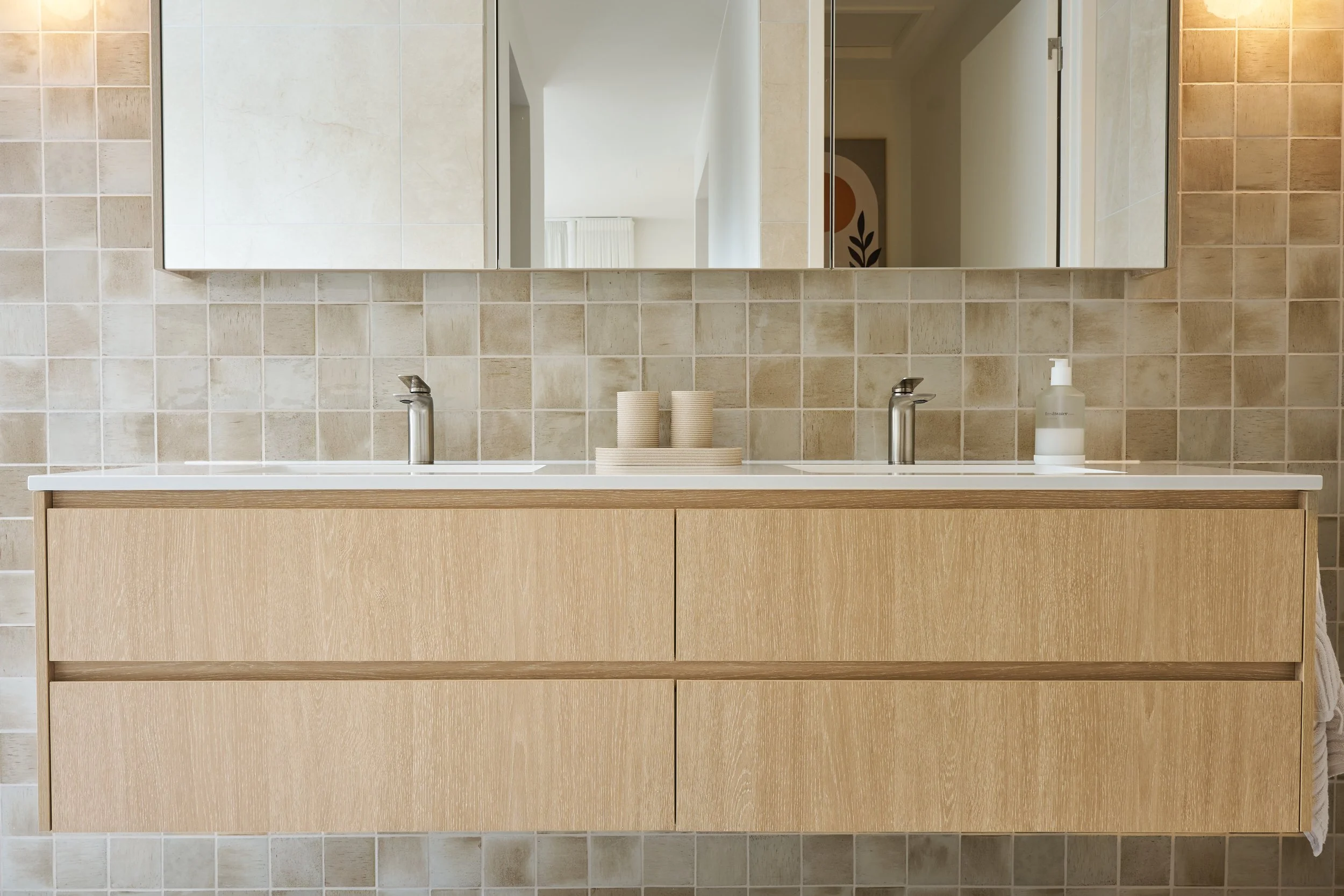 Overhead view of a modern bathroom vanity with a white countertop, two silver faucets, and a beige wooden cabinet below, mounted on a wall with beige square tiles. A mirror reflects part of the room and some beige cups and a soap dispenser are on the