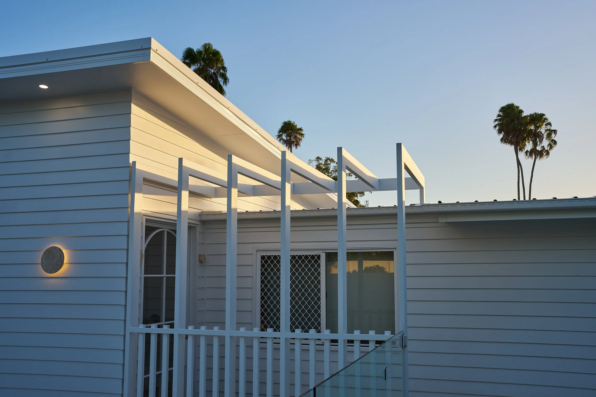 A modern white house with horizontal siding, a small balcony with white railing, and palm trees in the background under a clear blue sky.