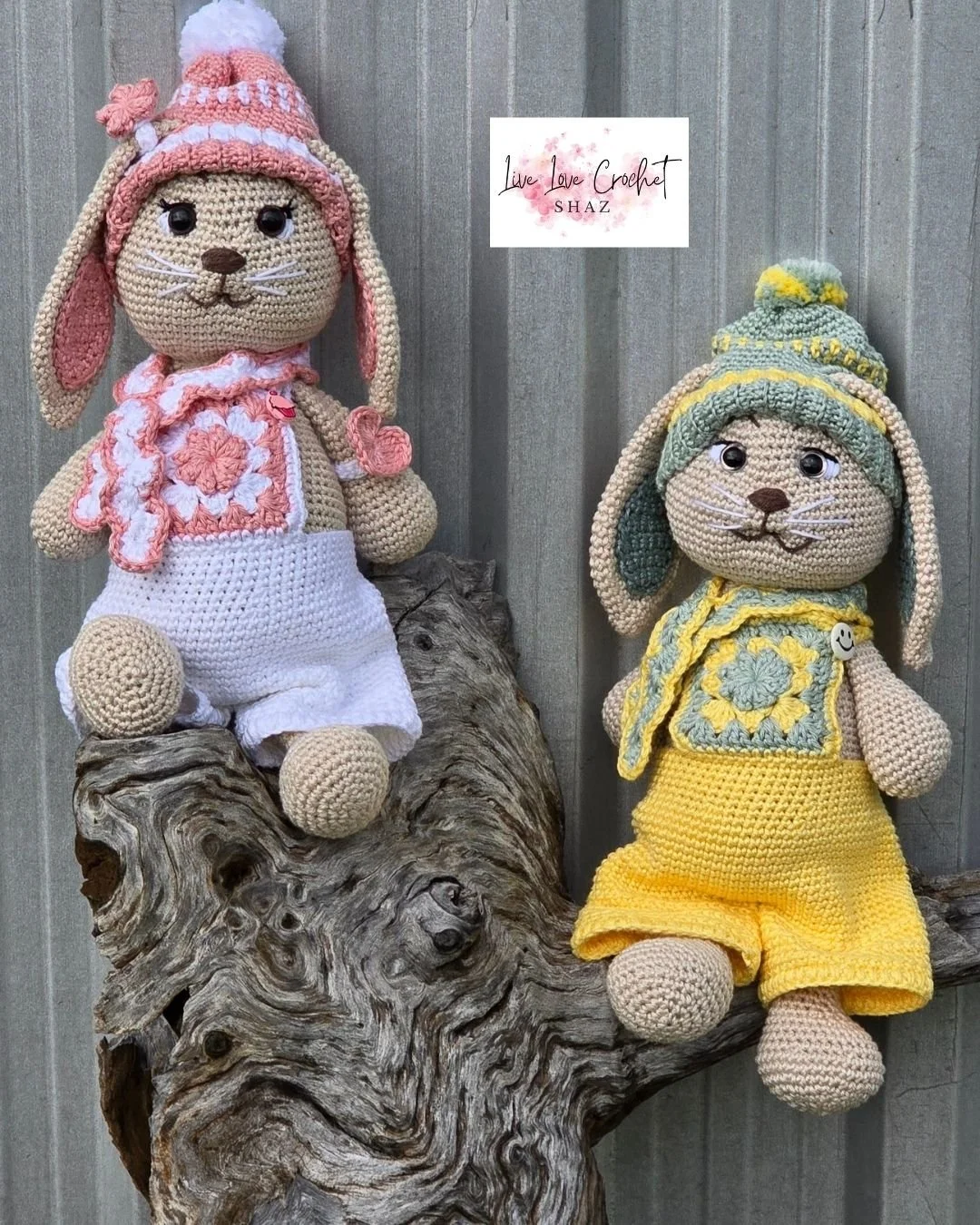 Two crocheted stuffed animal rabbits wearing colorful hats and clothing, sitting on a wooden branch in front of a gray wooden wall. One rabbit is dressed in pink and white, and the other in green and yellow. There is a sign that reads 'Live Love Crochet SHAZ' in the background.