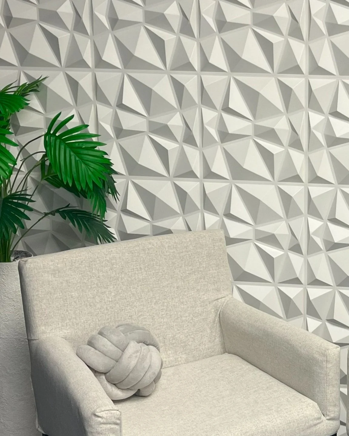 A beige sofa with a knotted cushion, a green plant with large leaves in a white pot, and a textured geometric white wall in the background.