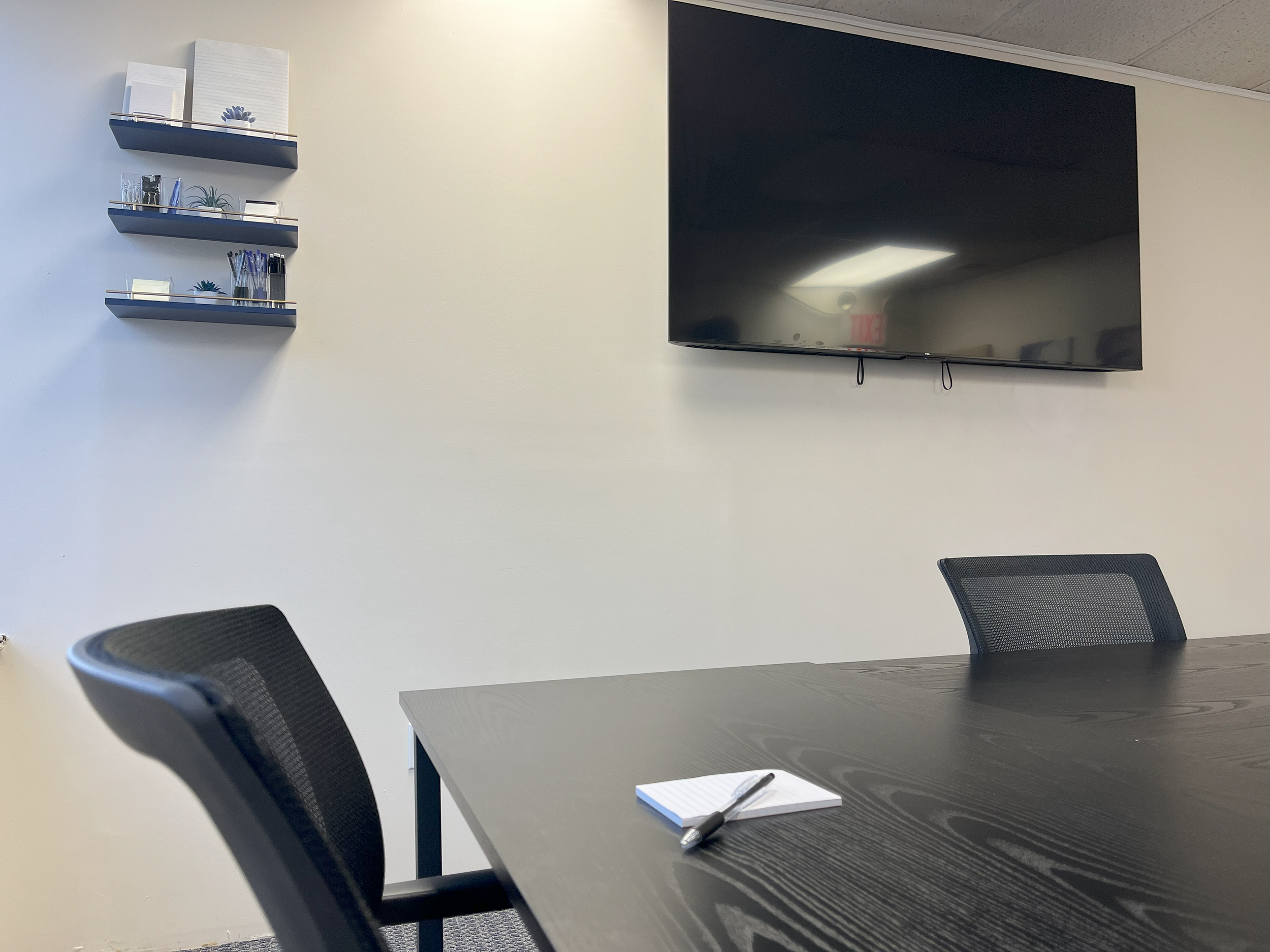 Empty conference room with black table, black chairs, wall-mounted TV, and shelves with office supplies.