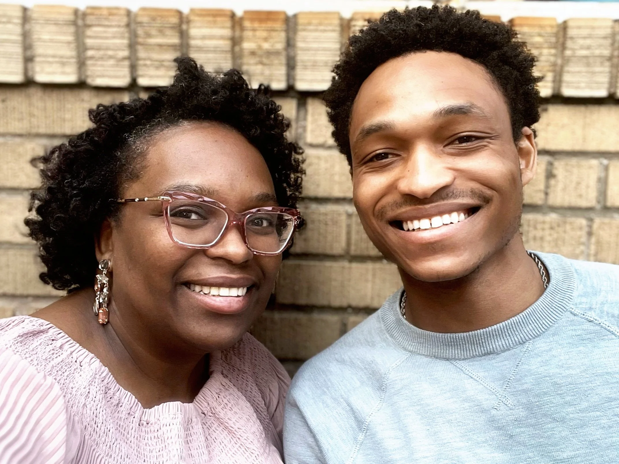 A smiling woman and a smiling man taking a selfie together in front of a brick wall.
