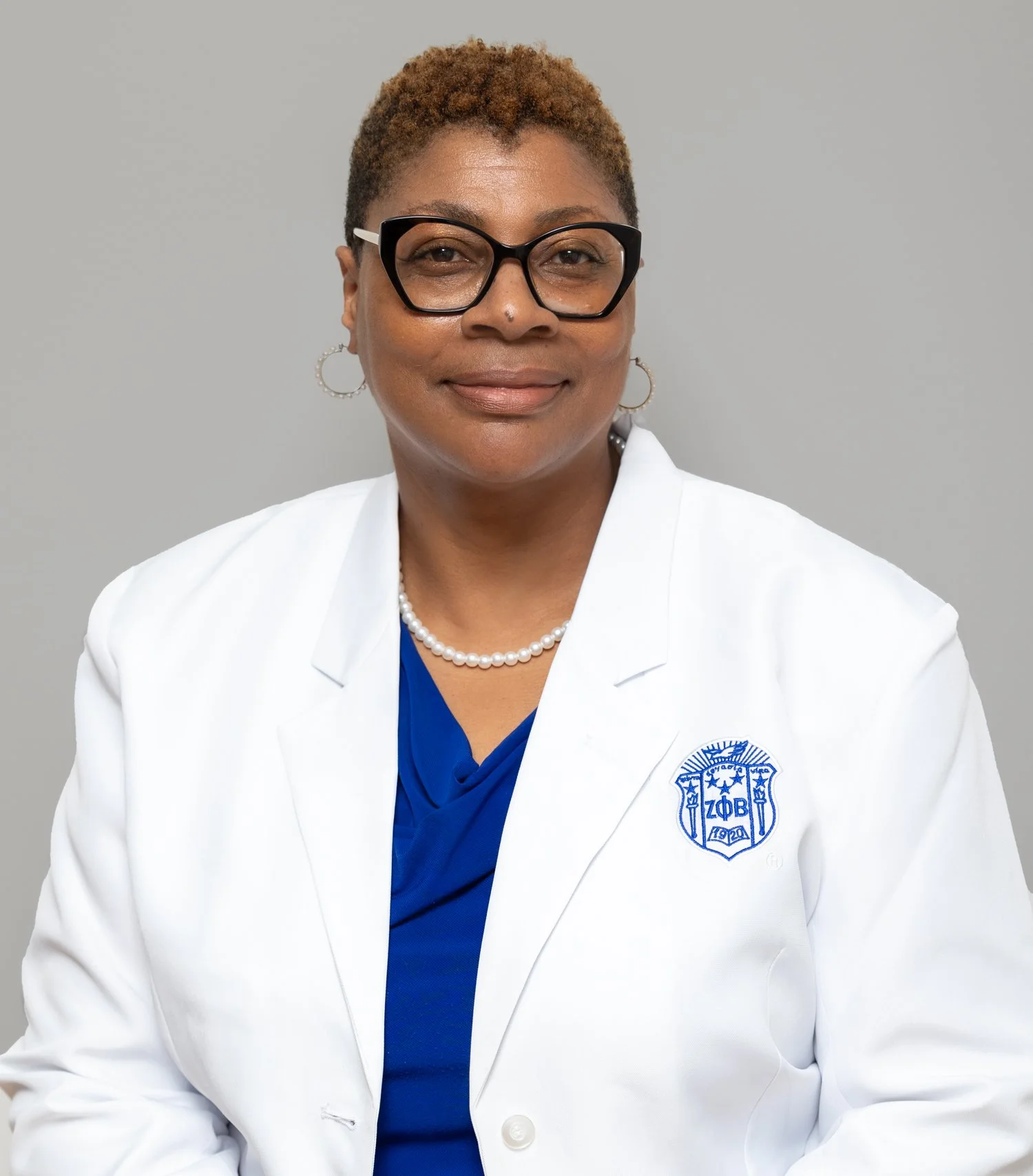 Portrait of an African American woman wearing a white lab coat with a blue shirt underneath, black glasses, pearl necklace, and hoop earrings, standing against a plain grey background.