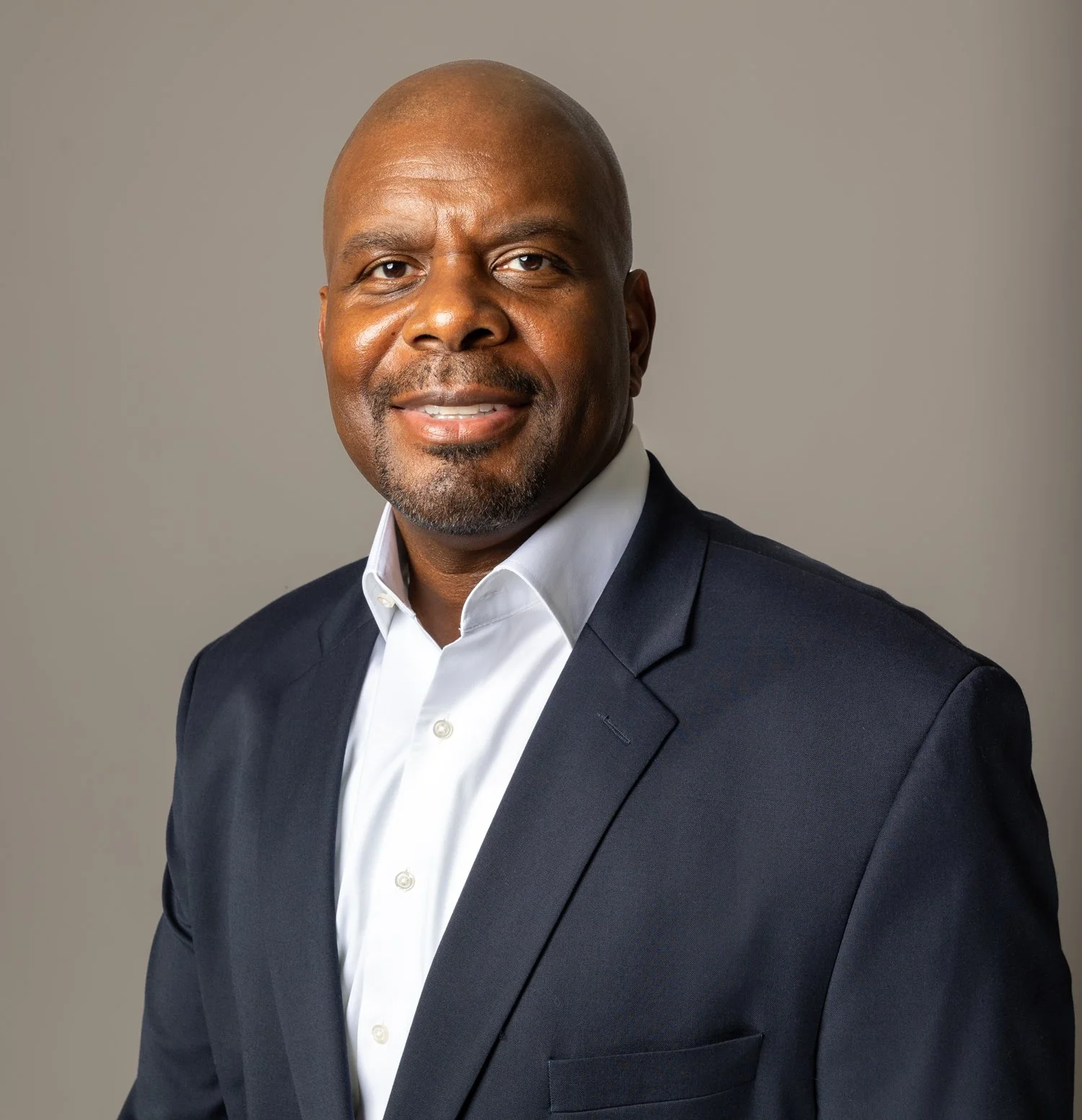 A professional headshot of a middle-aged African American man dressed in a dark suit jacket and white dress shirt, posing against a plain gray background.