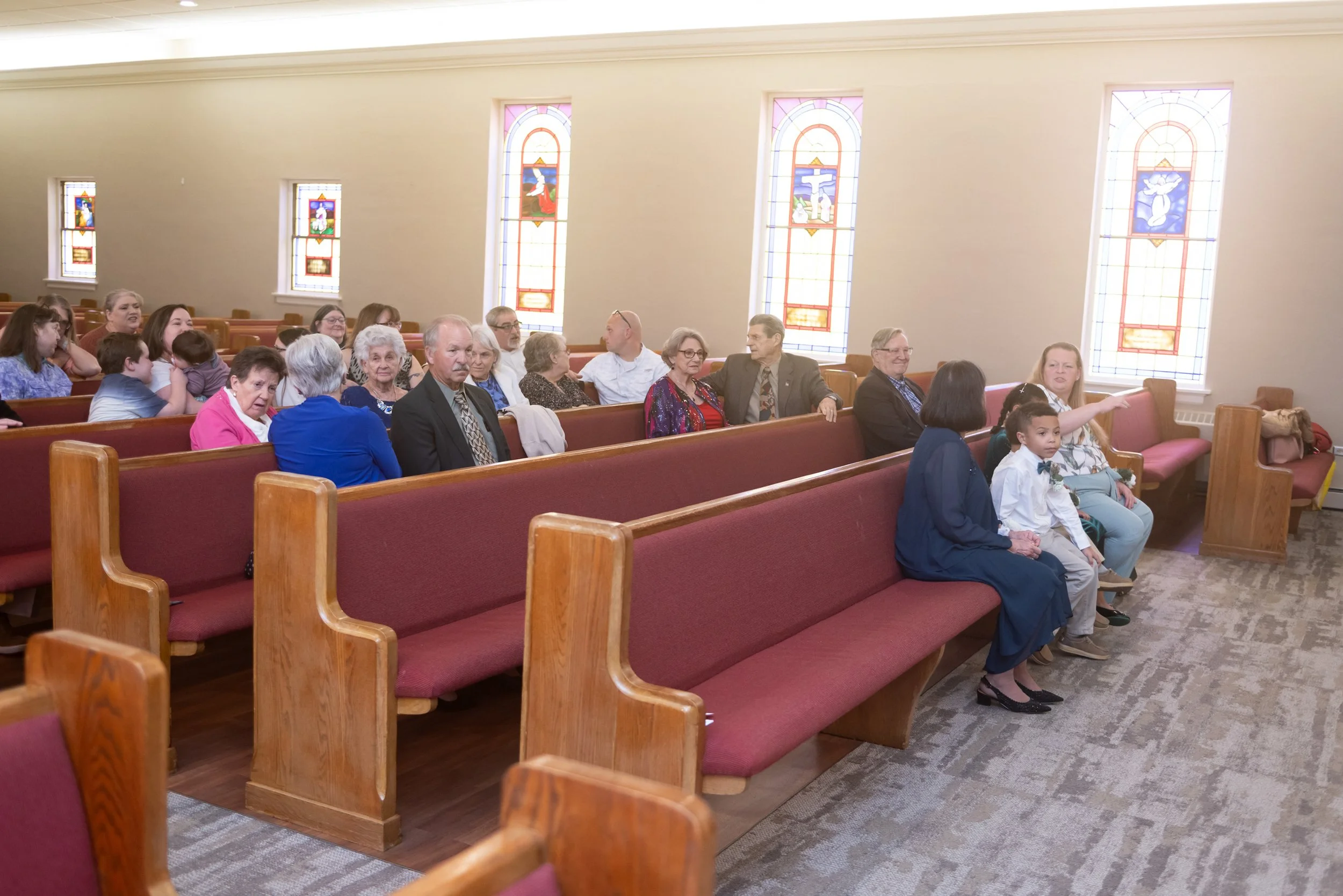 People sitting inside a church with stained glass windows, some engaging in conversation, and a woman pointing at something.