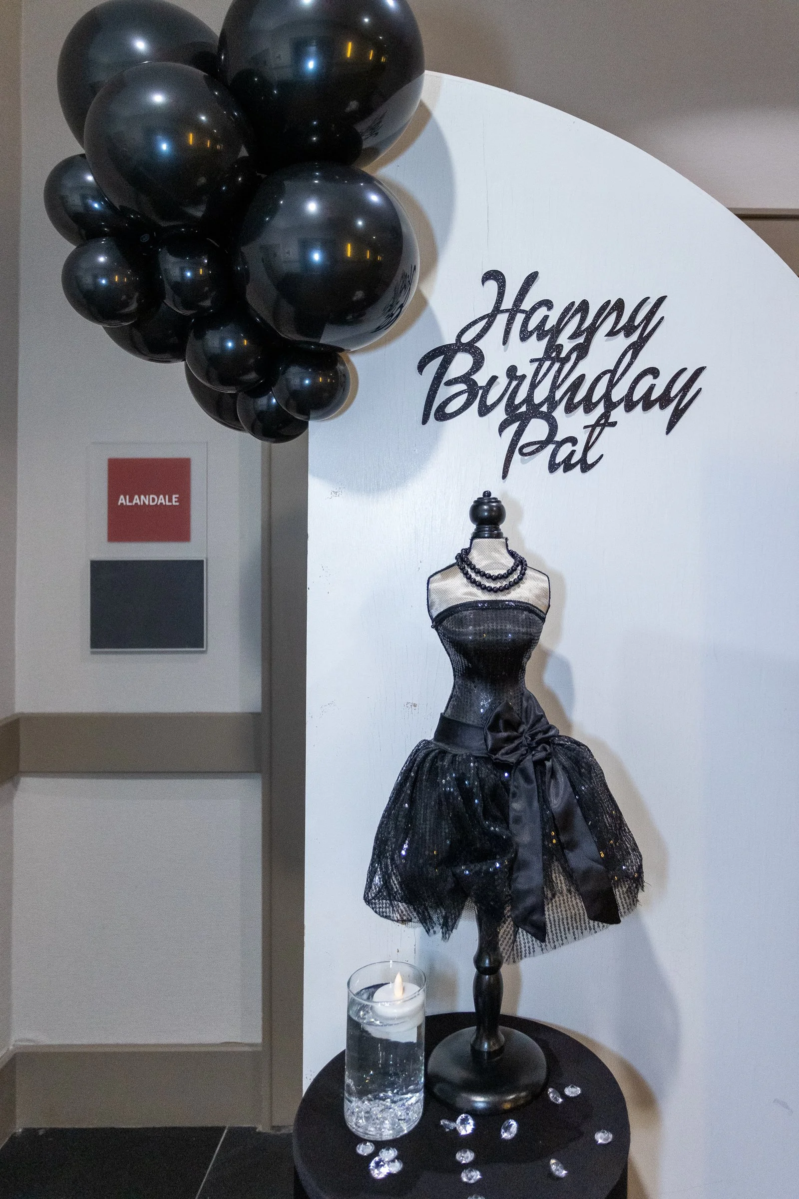 Black balloons clustered together, a white circular sign with black cursive text that reads 'Happy Birthday Pat', a black mannequin torso dressed in a black sequined dress with a large bow, a pearl necklace, and a black ribbon at the waist, a lit whi