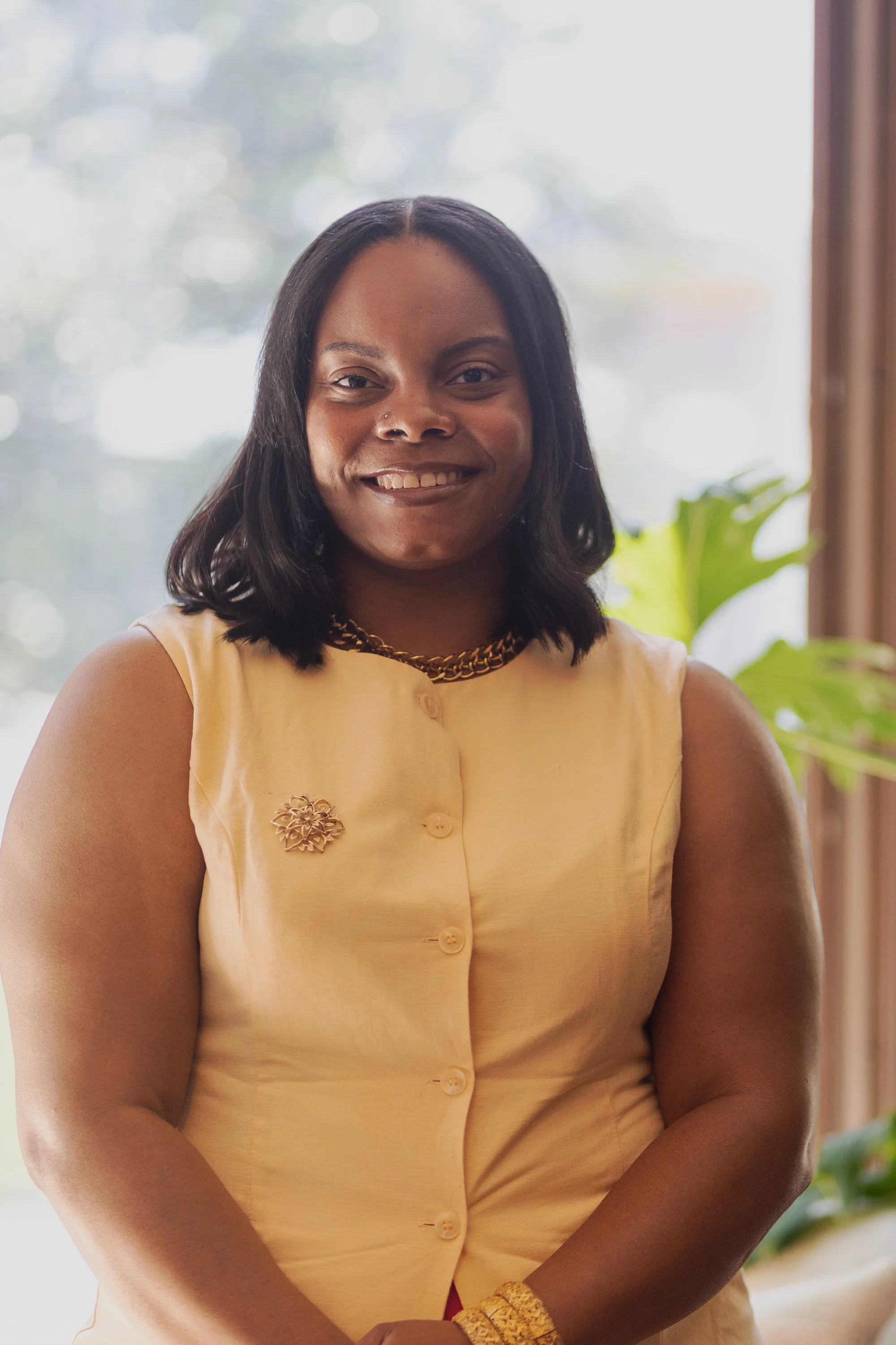 A woman smiling at the camera, wearing a yellow sleeveless top with a floral brooch, a gold chain necklace, and a yellow bracelet, standing indoors near a window with a blurred scenic background.