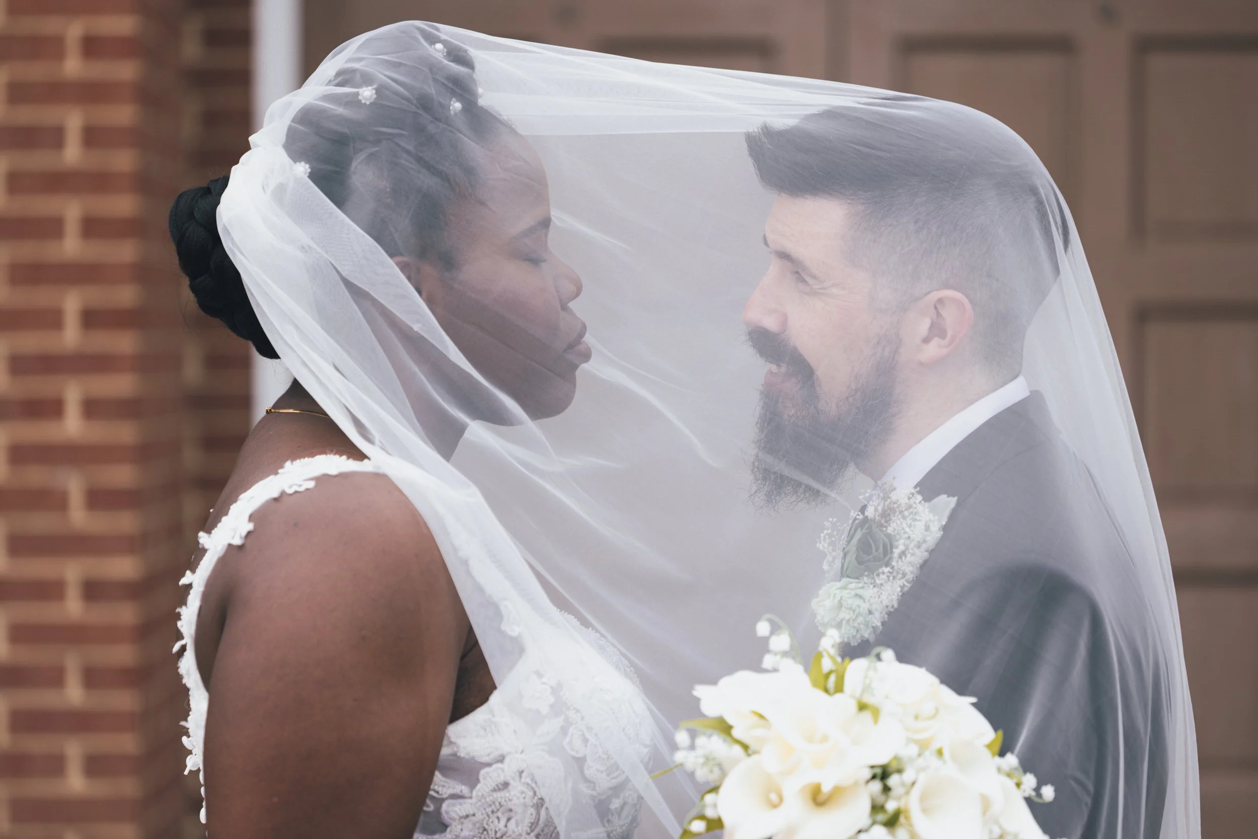 A bride and groom sharing a close moment under a wedding veil indoors in front of a brick wall.