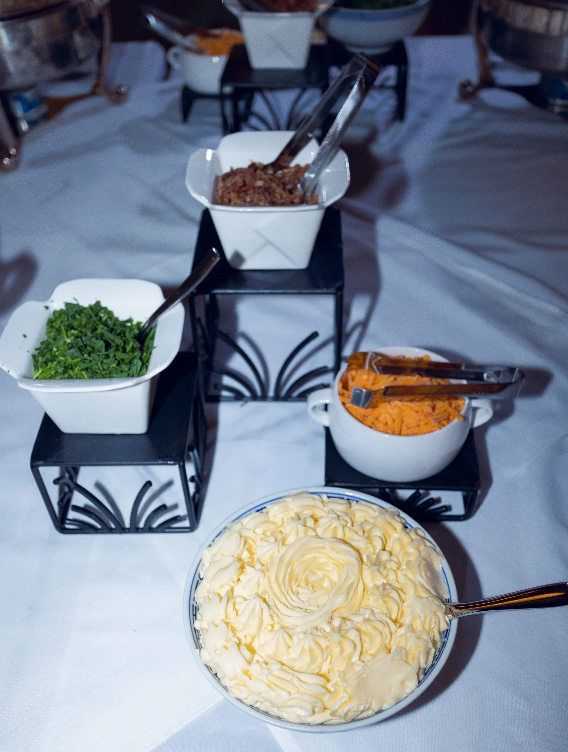 A large bowl of vanilla cake with buttercream frosting next to bowls of shredded cheese, chopped green herbs, shredded carrots, and a dish of minced meat, likely for a buffet or buffet-style meal.